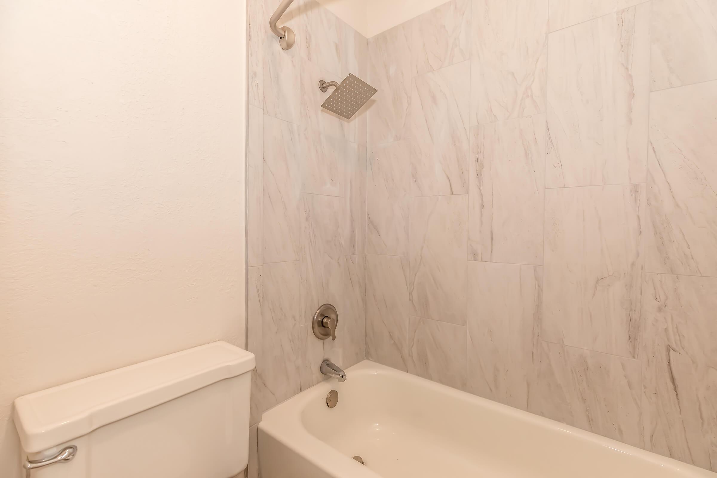 A clean bathroom featuring a bathtub with a showerhead above, white walls with a marble-like tile pattern, and a toilet beside the bathtub. The overall look is modern and minimalistic.