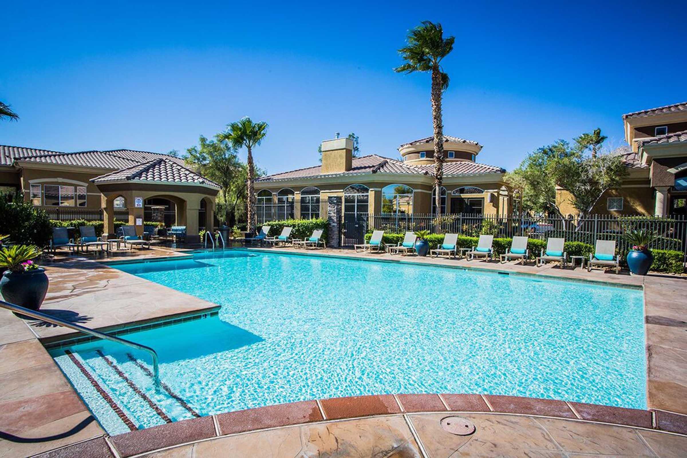 A sunny outdoor swimming pool surrounded by lounge chairs and palm trees. The pool features clear blue water and is situated near a stylish building with large windows. The scene conveys a relaxing and inviting atmosphere, perfect for leisure activities.