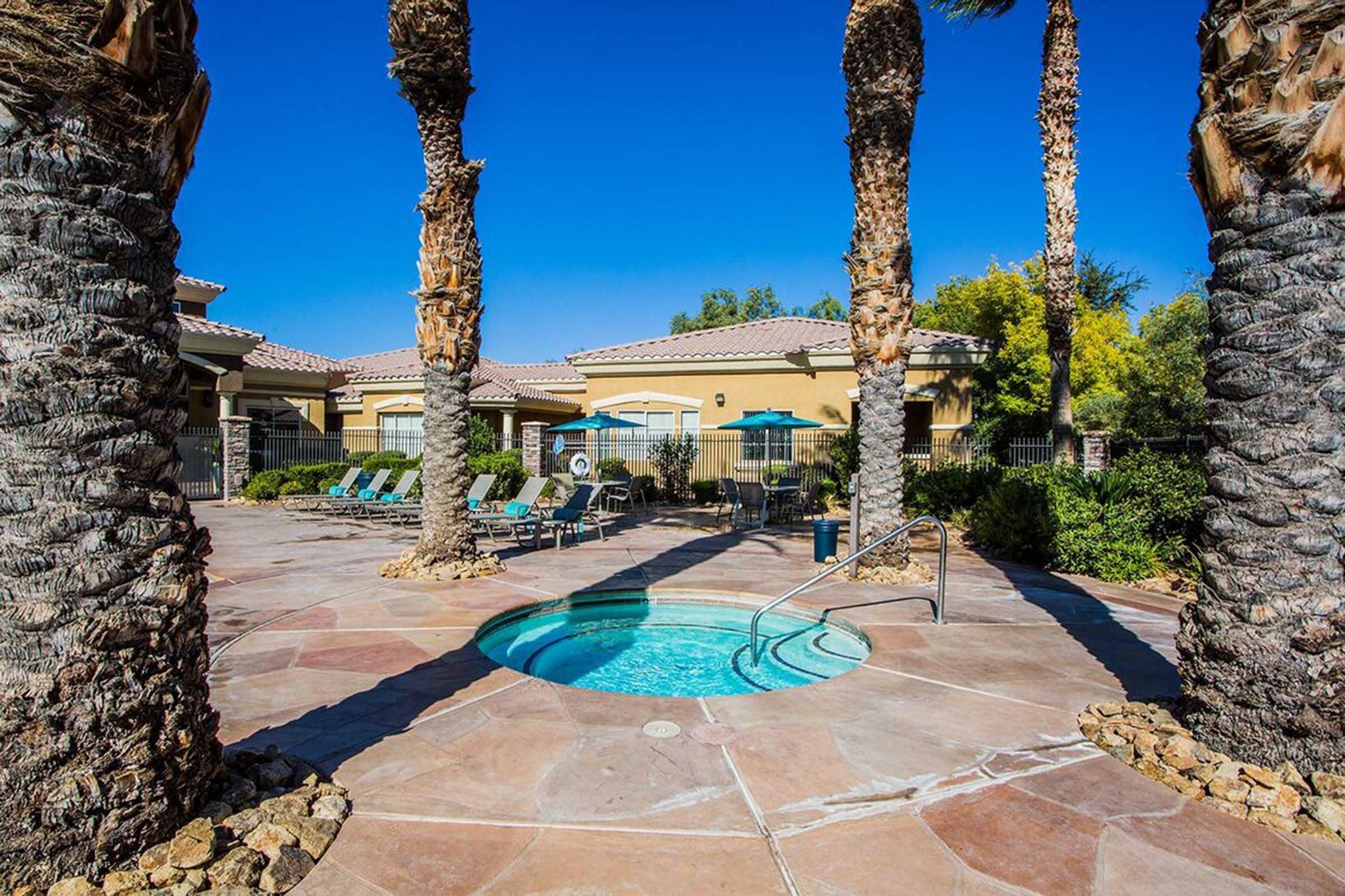 A sparkling hot tub surrounded by palm trees and lounge chairs in a sunny outdoor pool area. The backdrop features a well-maintained building with a blue sky overhead, creating a relaxing and inviting atmosphere.