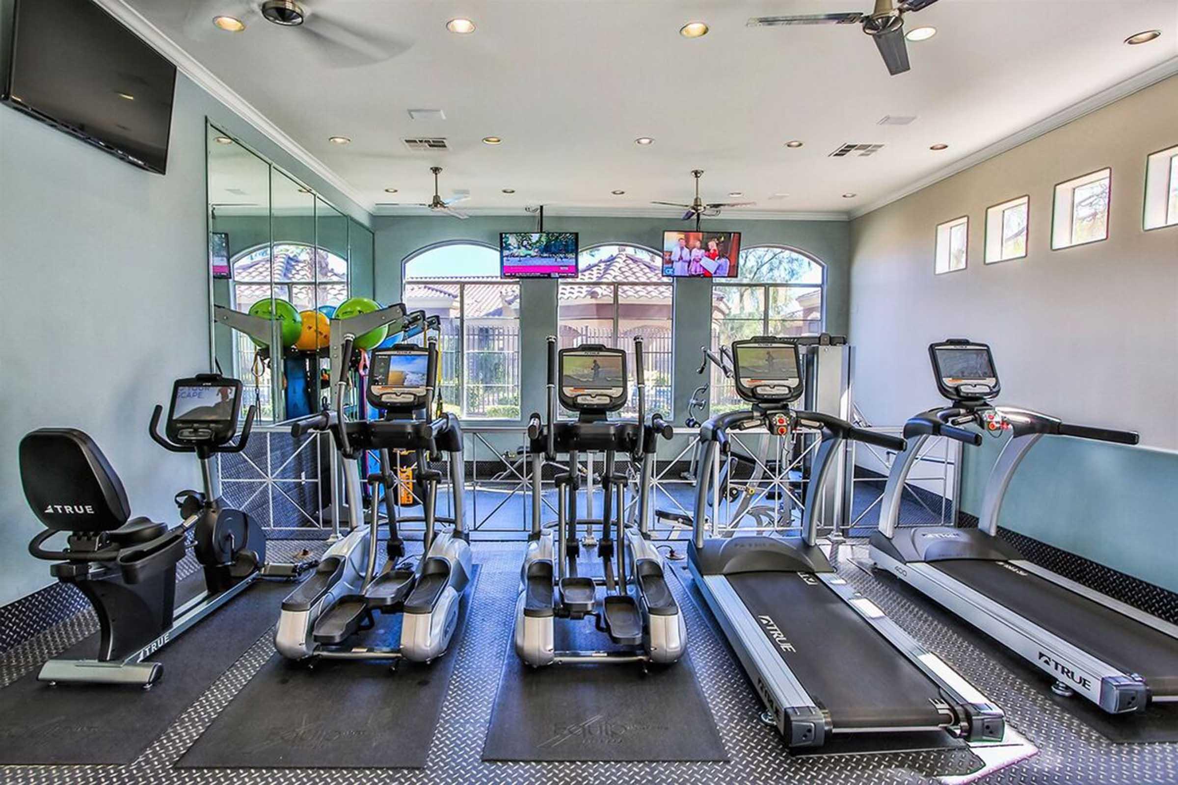 A modern gym area featuring multiple exercise machines including treadmills and elliptical machines. Large windows provide natural light, and there are TVs mounted on the walls. The floor is covered with black rubber mats for safety. Green exercise equipment can also be seen in the background.