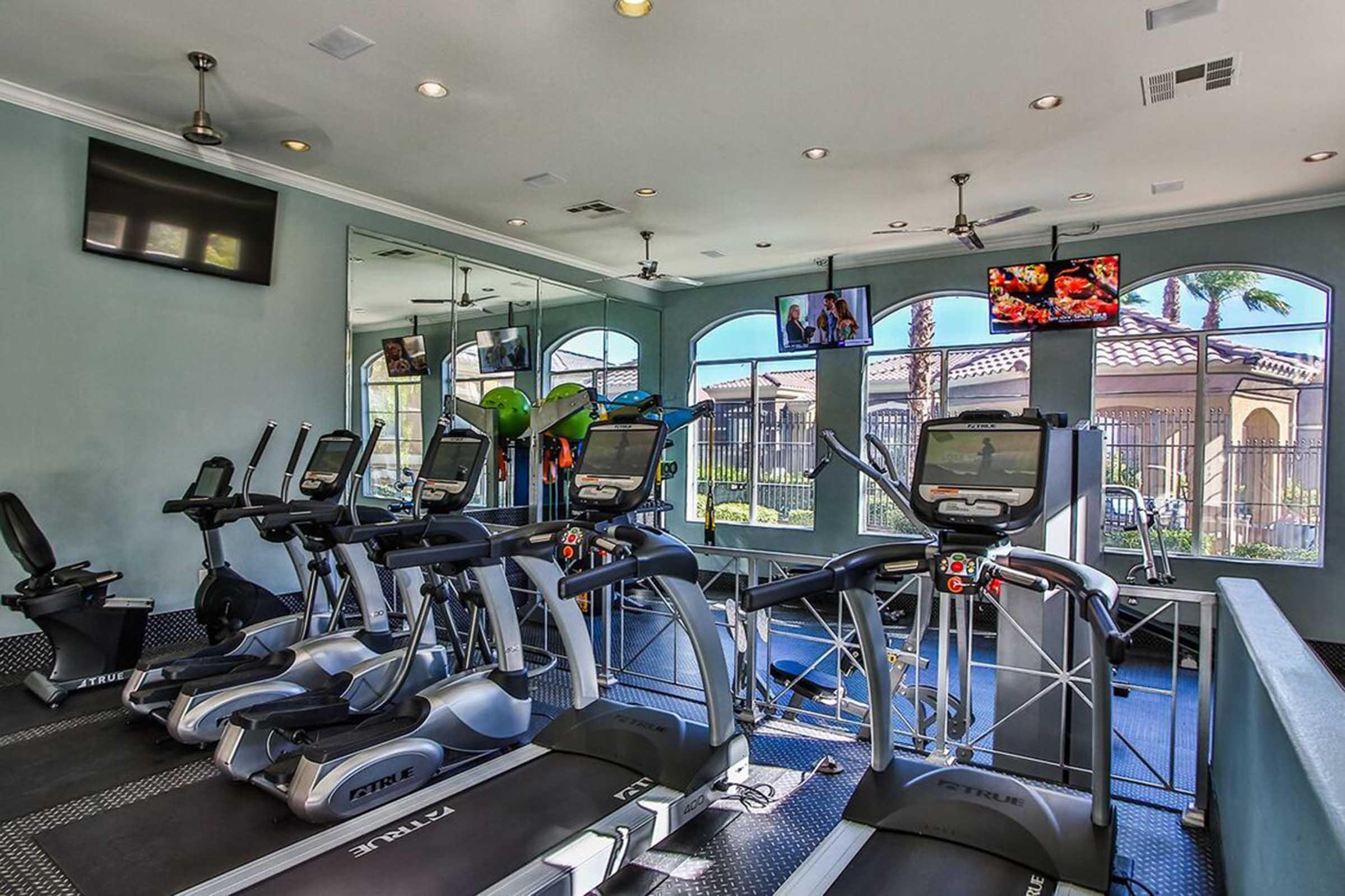 A modern gym interior featuring multiple treadmills and ellipticals arranged in a row. Large windows provide natural light and a view of the outdoors. Two televisions display content on the wall. The space is well-lit with a clean and inviting atmosphere, complete with plants for decoration.