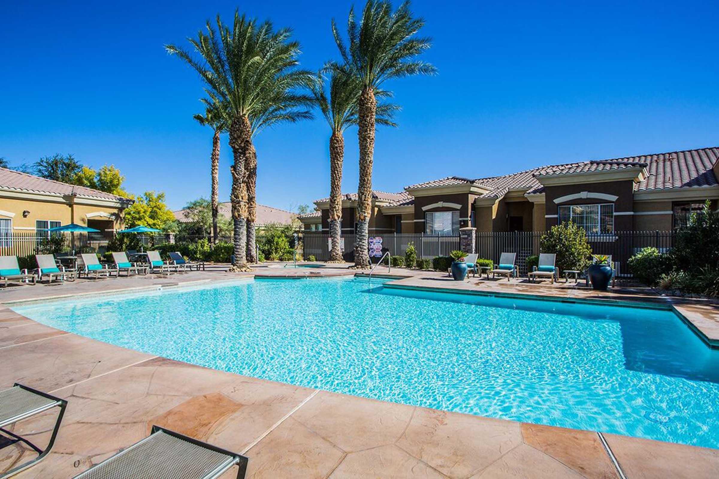 A serene swimming pool surrounded by palm trees and lounge chairs, set against a clear blue sky. In the background, there are well-maintained buildings and lush greenery, creating a relaxing outdoor atmosphere. Ideal for leisure and relaxation.