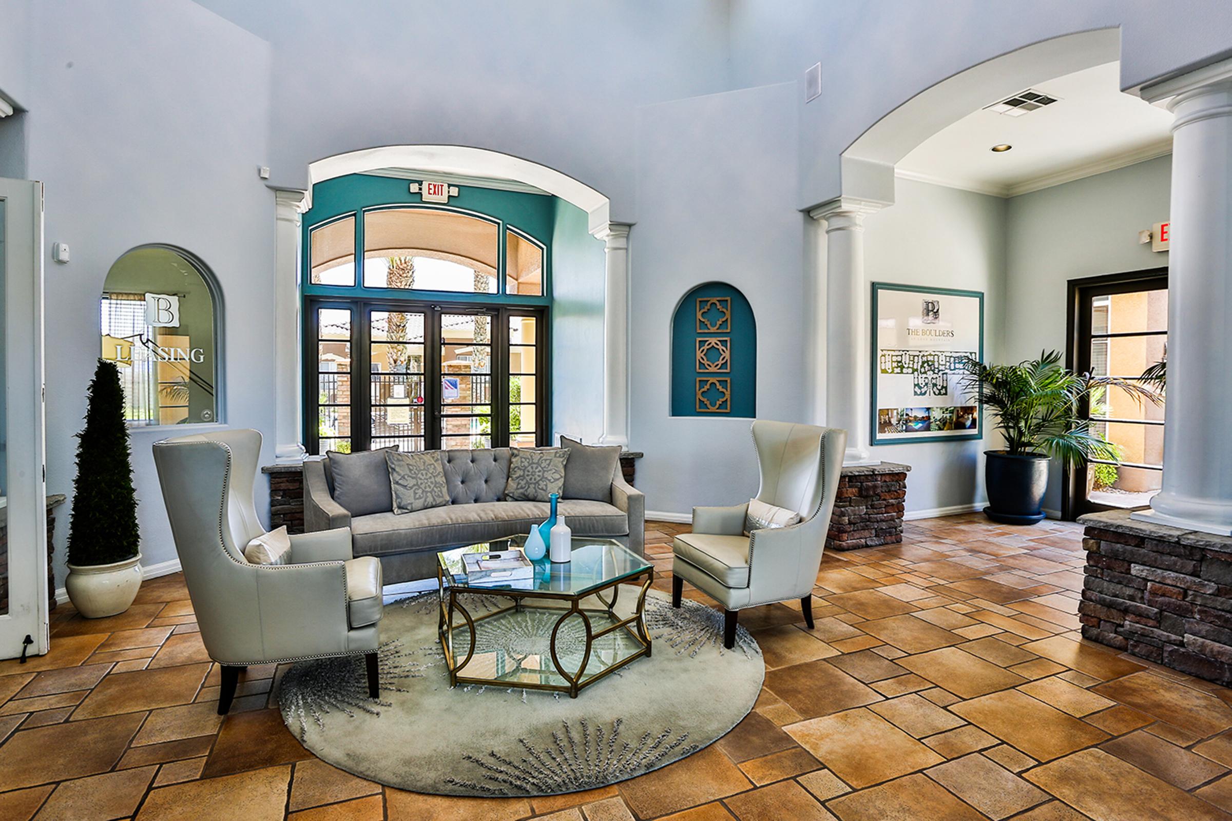 A modern lobby with light blue walls features stylish seating arrangements, including a gray sofa and armchairs around a glass coffee table. Decorative elements include plants, artwork, and a geometric rug. Large windows allow natural light, creating a welcoming atmosphere.