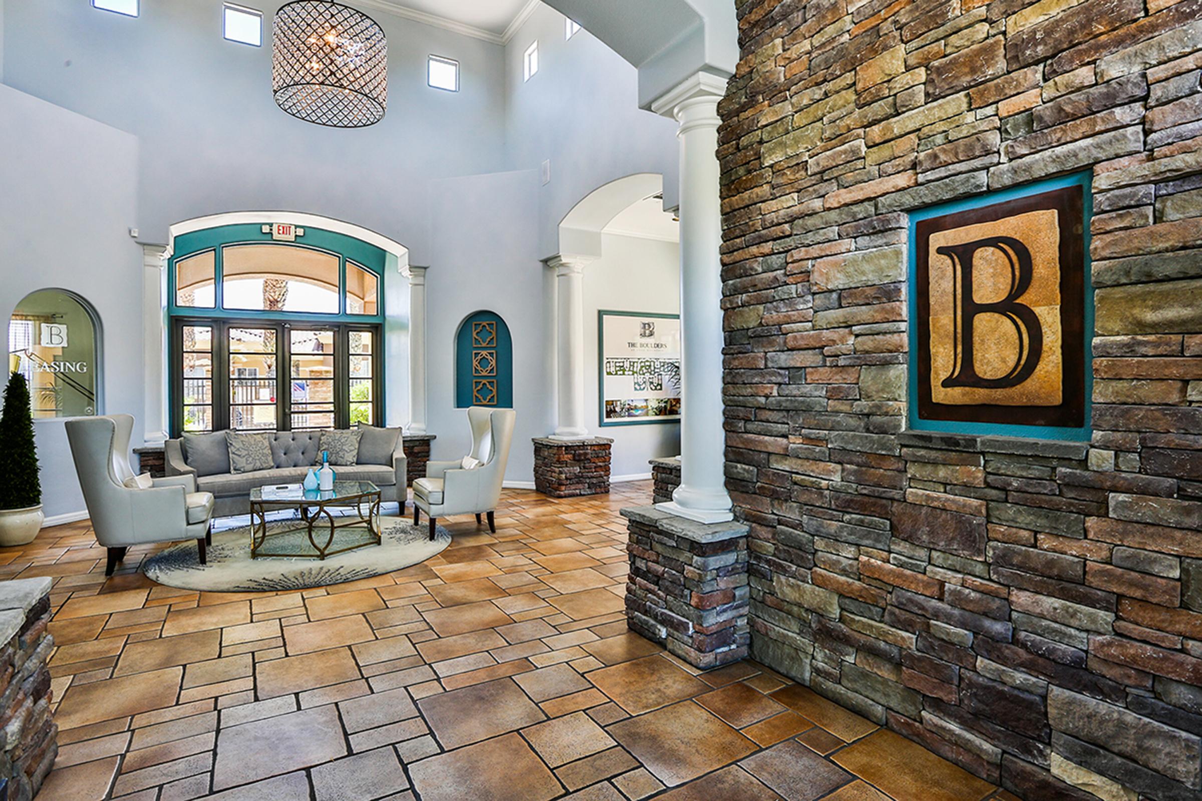 A spacious lobby with a high ceiling, featuring a chandelier and large windows. The floor is tiled, and there are stone accent walls. Comfortable seating is arranged in the center, with a round rug and a small table. The wall on the right displays a large letter "B" in a decorative frame.