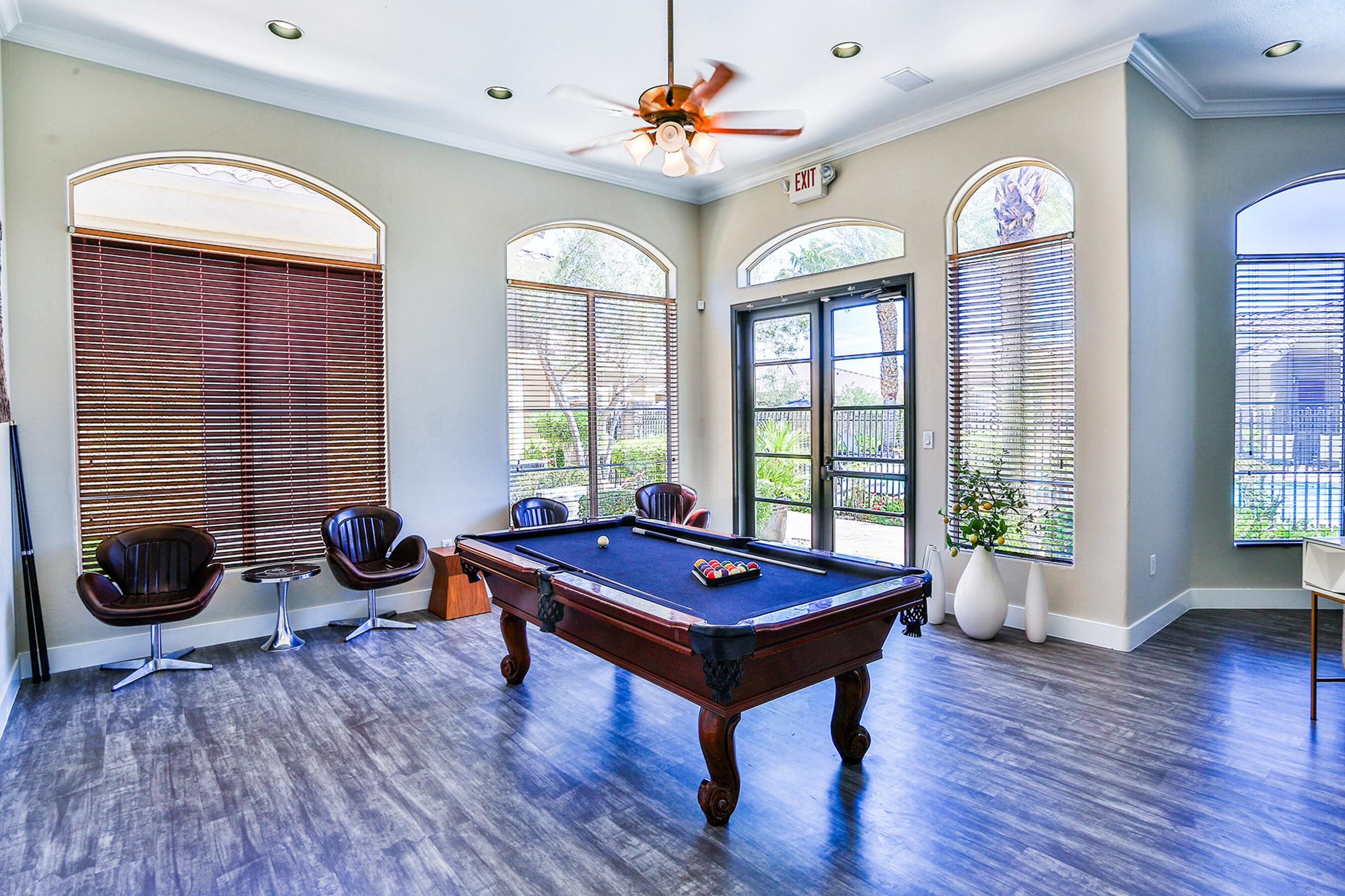 A bright indoor space featuring a pool table with a set of colorful balls on it. There are large windows with wooden blinds, allowing natural light to flood the room. Stylish chairs are arranged nearby, and potted plants add a touch of greenery to the decor.