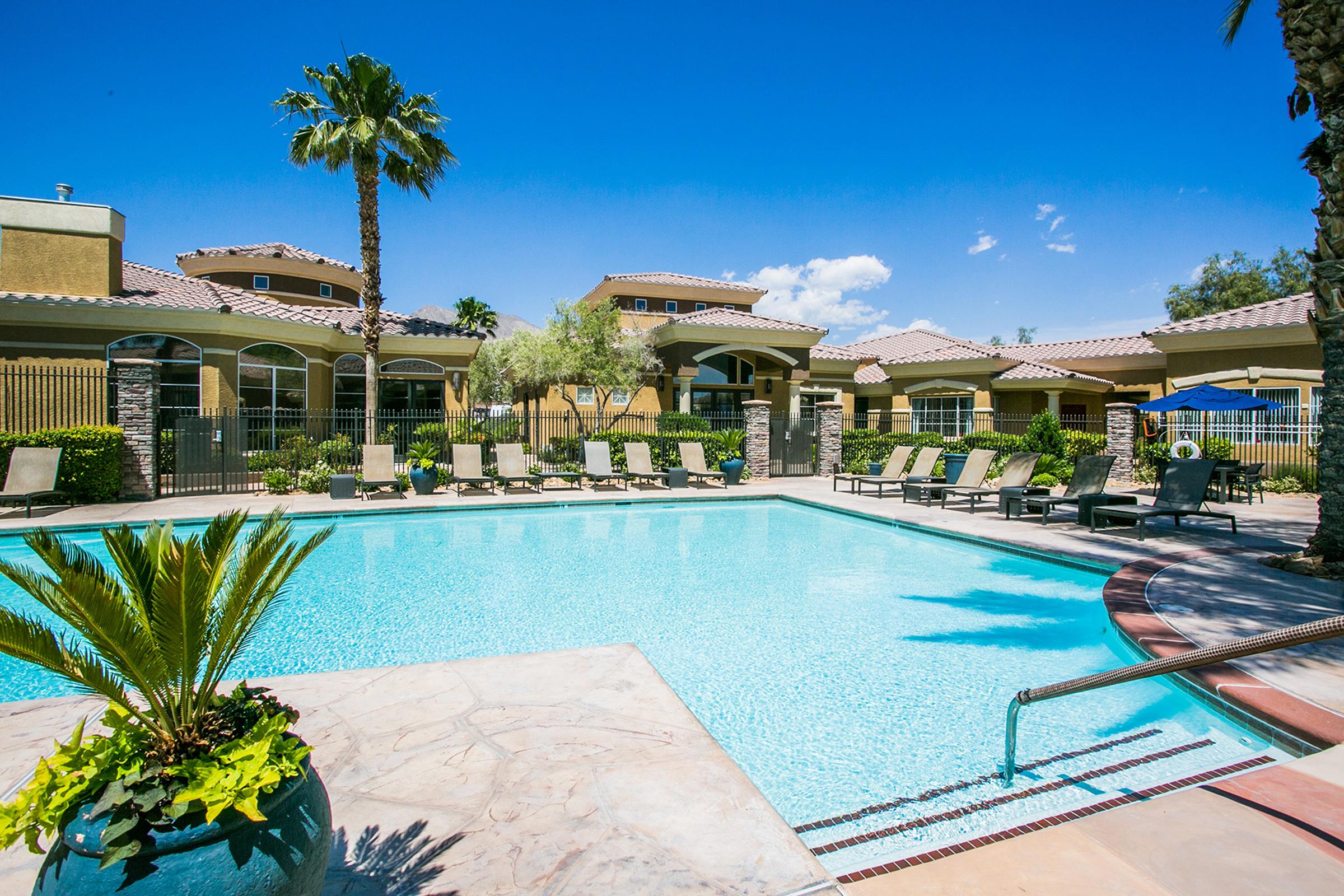 A sunny pool area featuring a clear blue swimming pool surrounded by lounge chairs, palm trees, and landscaped greenery. In the background, there are residential buildings with a warm color scheme and a clear blue sky.