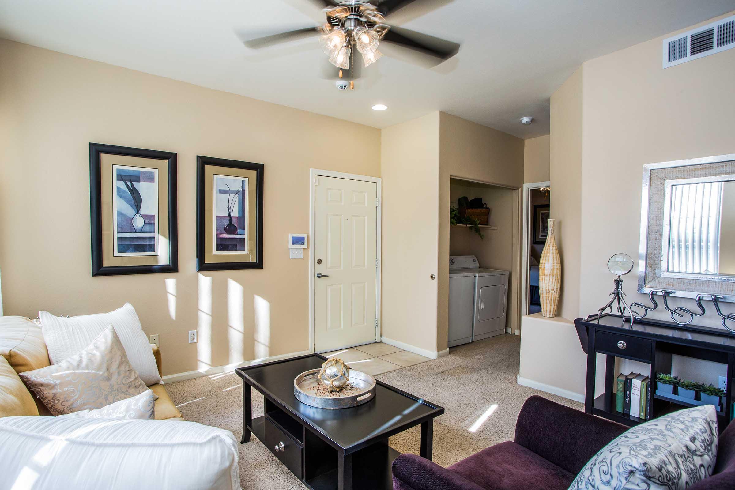 A cozy living room featuring a light-colored sofa, decorative pillows, and a dark coffee table with a circular centerpiece. Two framed artworks are hung on the wall, and a doorway leads to a laundry area. Natural light streams in through a window, creating a warm and inviting atmosphere.