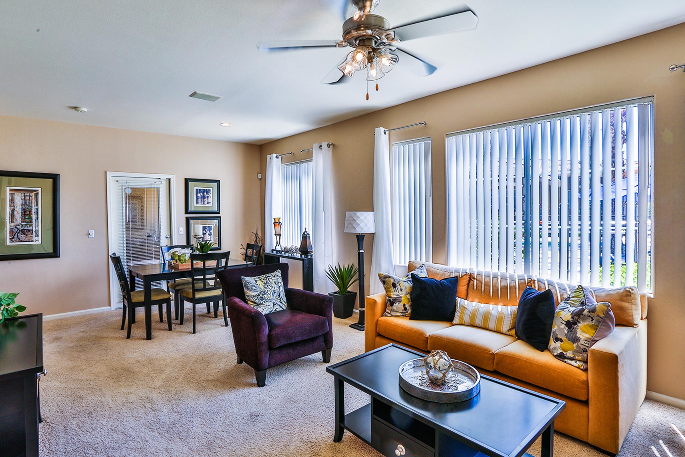 A cozy living room featuring a mustard yellow couch with patterned pillows, a dark coffee table, and an armchair. Natural light streams in through large windows with vertical blinds. A dining area with a table and chairs is visible, along with decorative plants and wall art, creating an inviting atmosphere.
