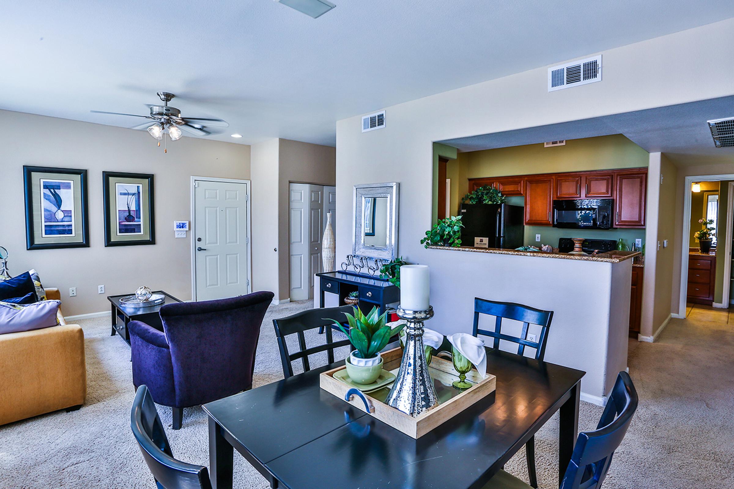 A bright and spacious apartment interior featuring a dining table with decorative items, a cozy living area with sofas and a ceiling fan, and an open kitchen with wood cabinetry in the background. Light-colored walls and carpeting create a warm atmosphere.