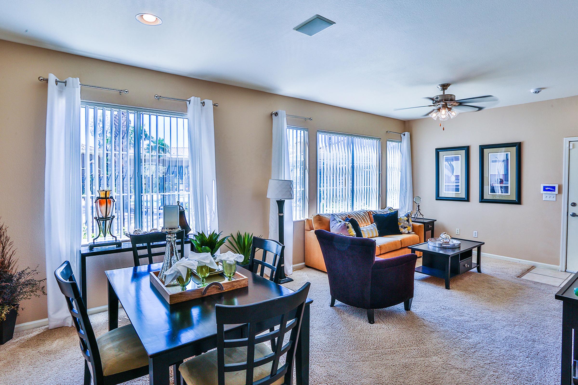 A spacious living room with large windows allowing natural light, featuring a cozy orange sofa, a dining table set for four, and decorative plants. The walls are painted a soft beige, and there are two framed pictures hanging near the windows. A ceiling fan is installed above, adding a touch of comfort.
