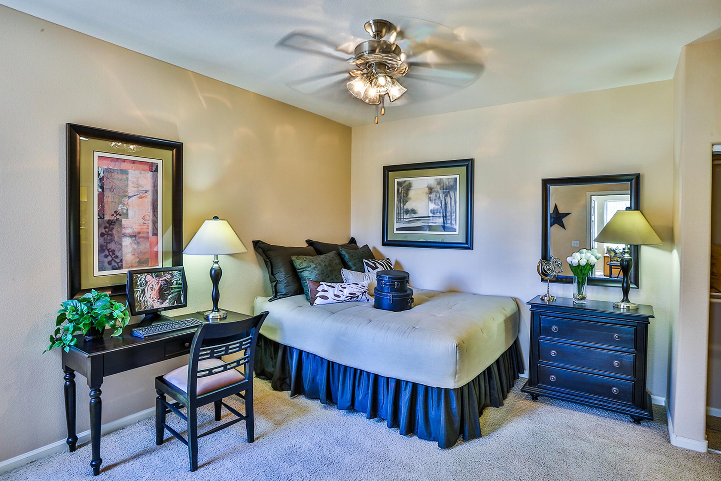 Cozy bedroom featuring a bed with decorative pillows, a ceiling fan, a desk with a chair, and a black nightstand. Two lamps provide soft lighting, and framed artwork adorns the walls. A small plant adds a touch of greenery, creating a welcoming atmosphere.
