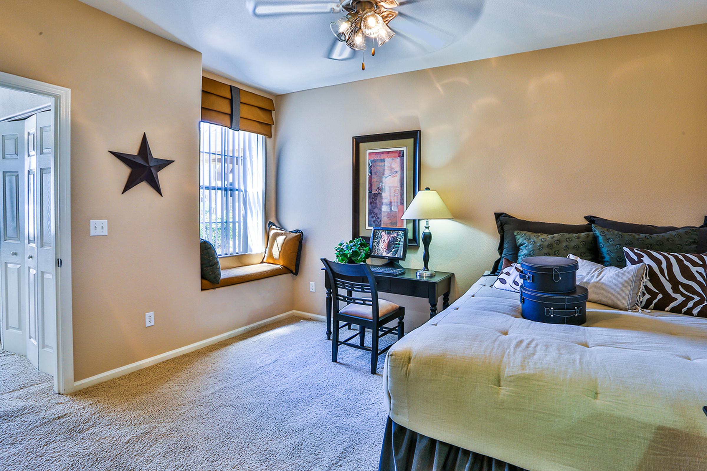 A well-lit bedroom featuring beige walls and carpet. A cozy window seat with decorative pillows is visible, along with a small desk and chair. A bedside table holds a lamp, and a large bed with patterned pillows is positioned in the corner. A decorative star hangs on the wall.