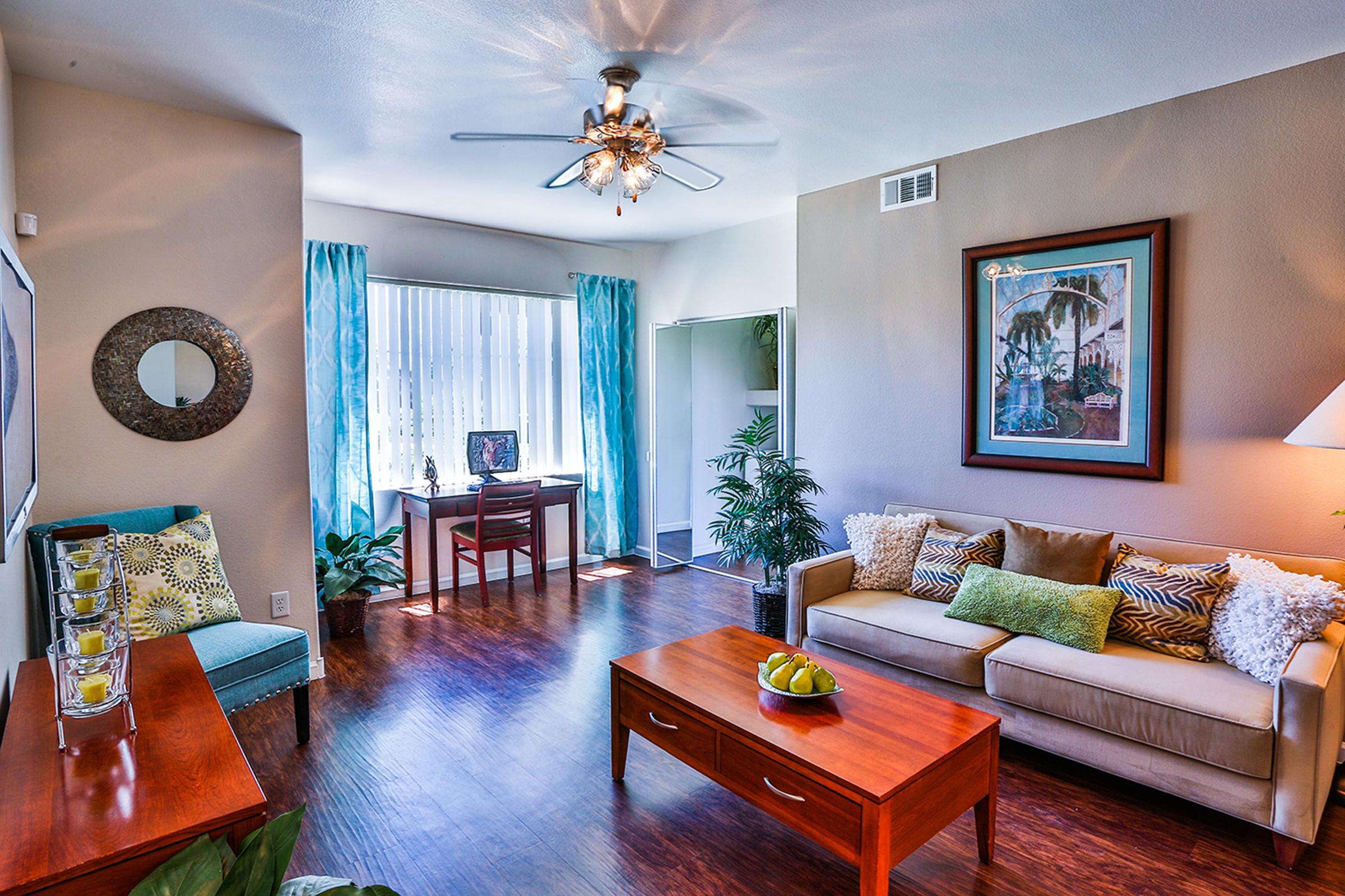 A modern living room featuring a beige sofa with colorful cushions, a wooden coffee table with decorative items, and a small workspace with a desk near a window. The walls are adorned with a framed artwork, and there are plants and decorative elements adding a vibrant touch to the space.