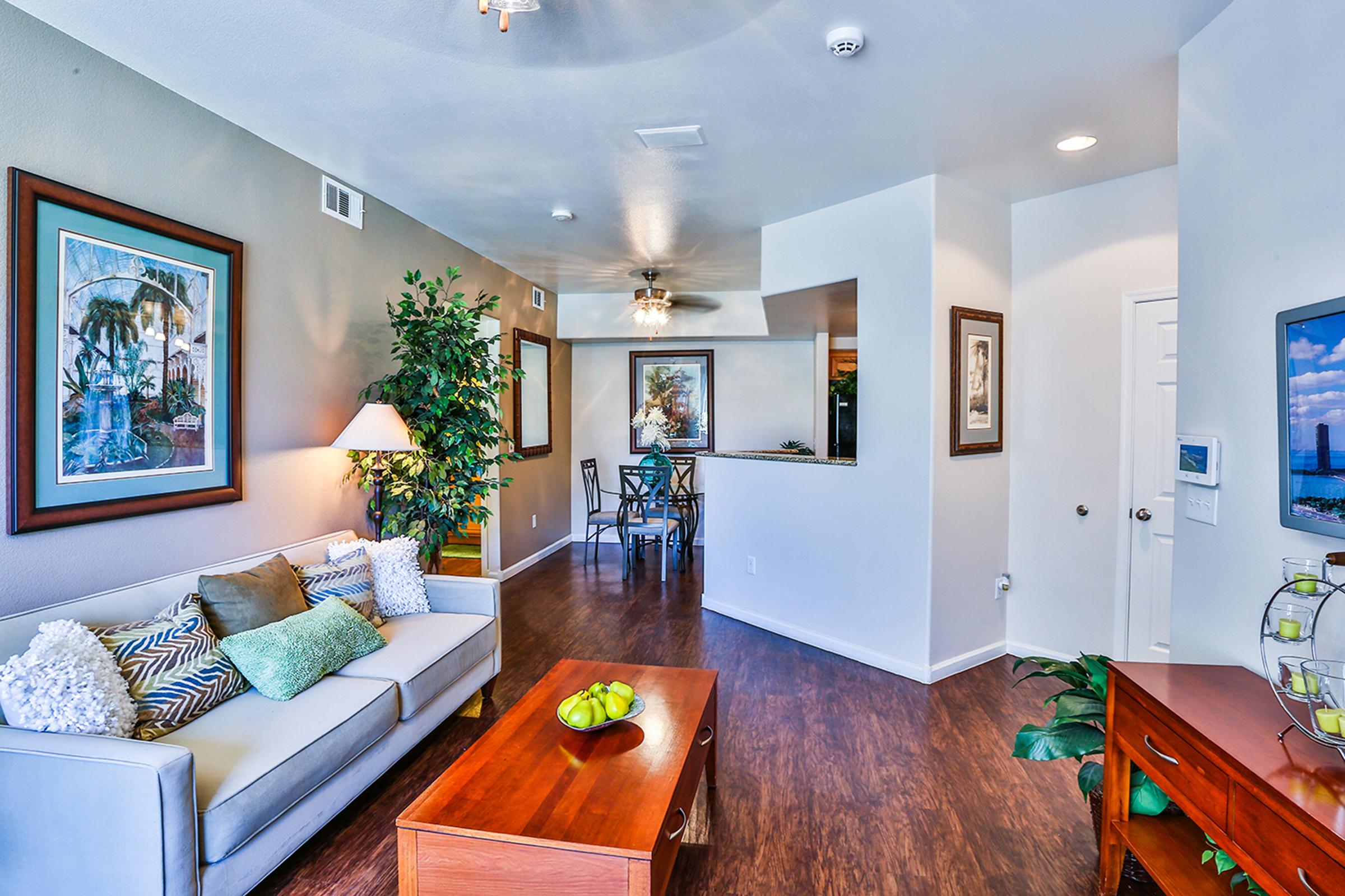 A stylish living room featuring a gray sofa with colorful pillows, a wooden coffee table with a bowl of green apples, and a decorative plant. The walls are adorned with art, and there’s an open layout leading to a kitchen area visible in the background. Natural light fills the space, creating a welcoming atmosphere.