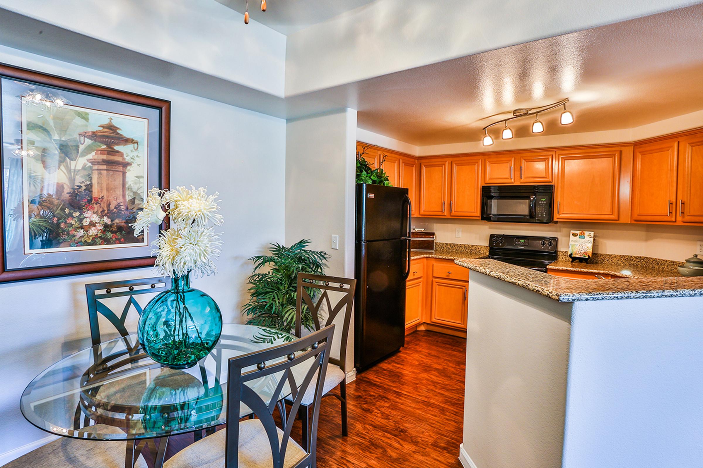 A modern kitchen and dining area featuring wooden cabinets, a granite countertop, and a glass dining table with decorative flowers. The space is well-lit, showcasing a black refrigerator and other kitchen appliances, with a framed artwork on the wall and plants adding a touch of greenery.