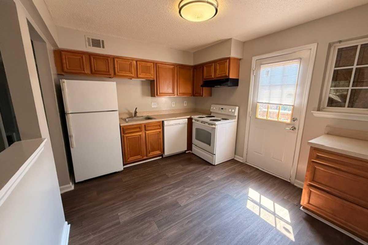 A bright, clean kitchen featuring light-colored walls, wooden cabinets, and modern appliances. There's a white refrigerator, dishwasher, and stove. A window allows natural light, and a door leads outside. The floor is dark vinyl, creating a contemporary look in the space.