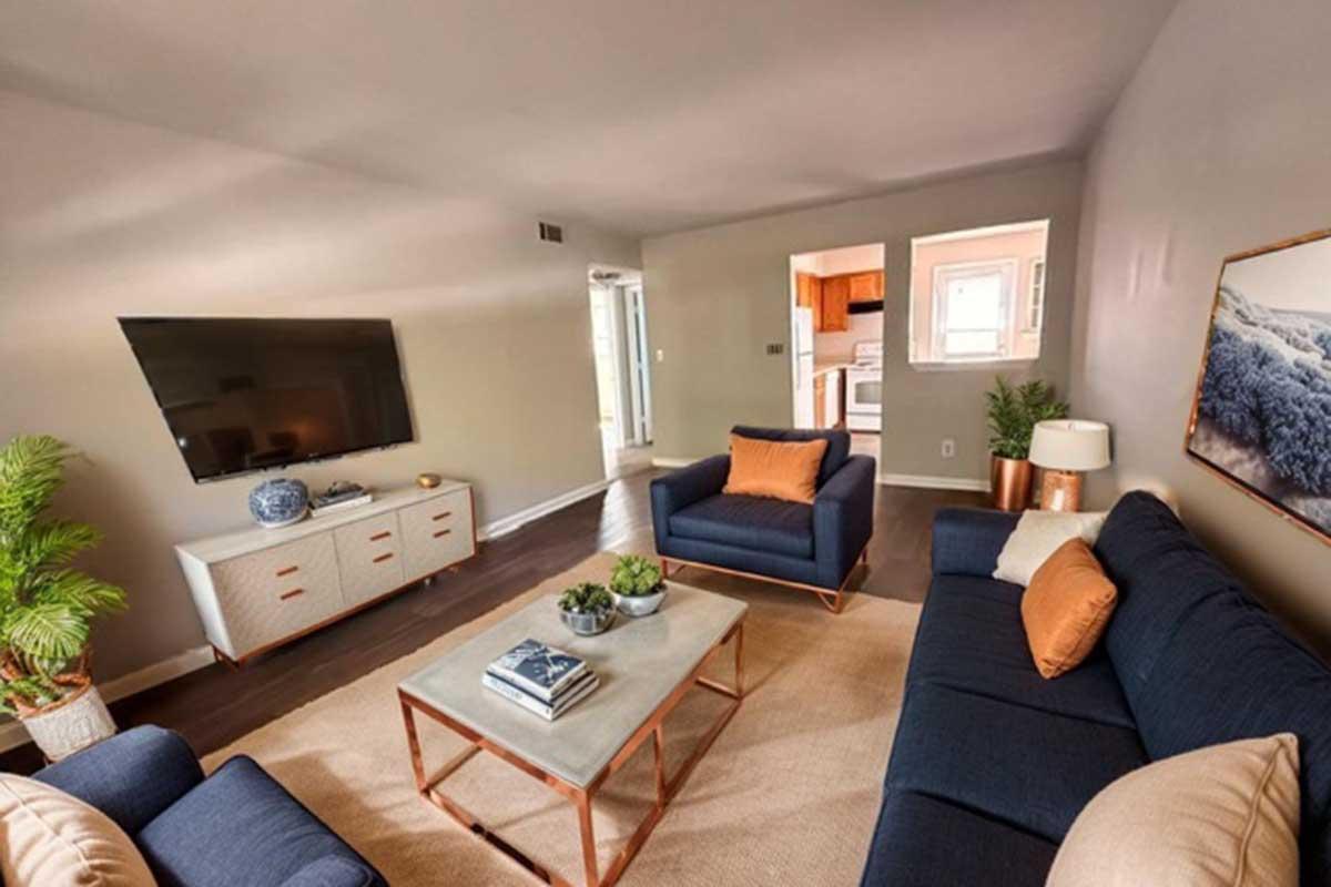 A modern living room featuring two navy blue sofas with orange cushions, a glass coffee table, and a light-colored area rug. There is a TV mounted on the wall, a console table underneath, and decorative plants. Natural light enters from a nearby window, illuminating the space.