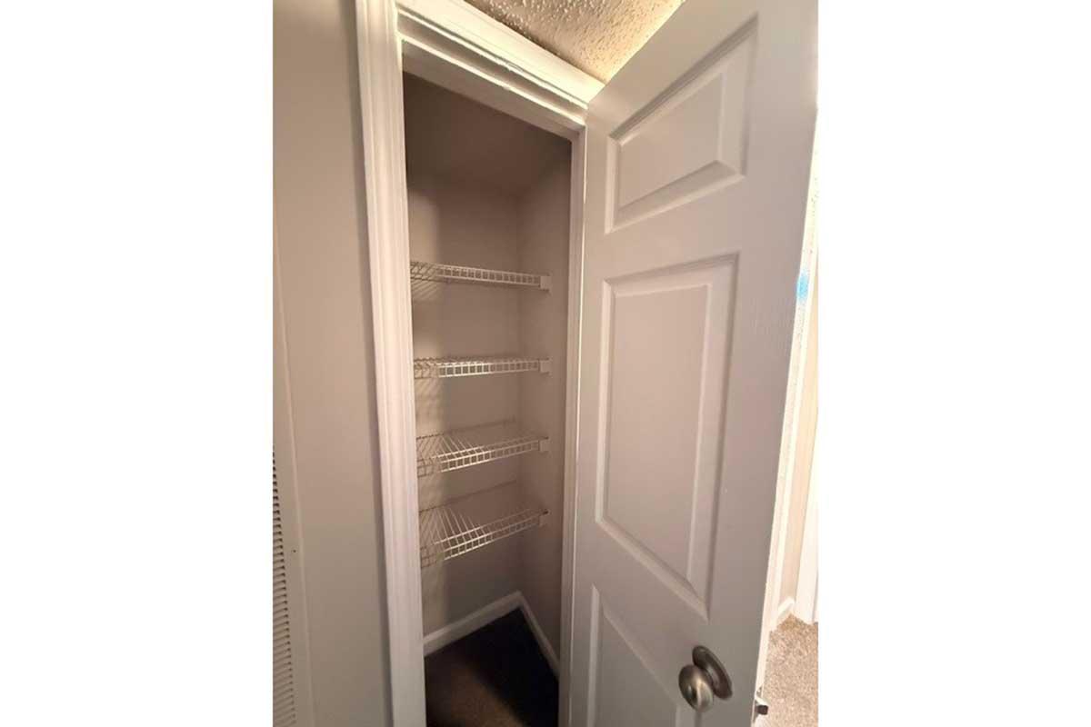 A view of an empty closet with white wire shelving. The closet has a white door that is slightly ajar, revealing the interior which is clean and well-lit. The walls and shelves are also white, creating a bright and spacious appearance.