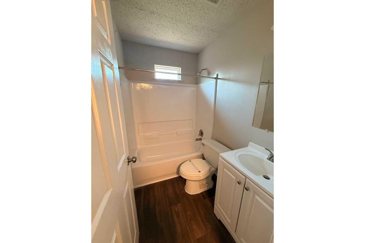A small bathroom featuring a white bathtub and shower, a toilet, and a sink with a cabinet. The walls are painted light gray, and there's a small window providing natural light. The flooring is dark wood.