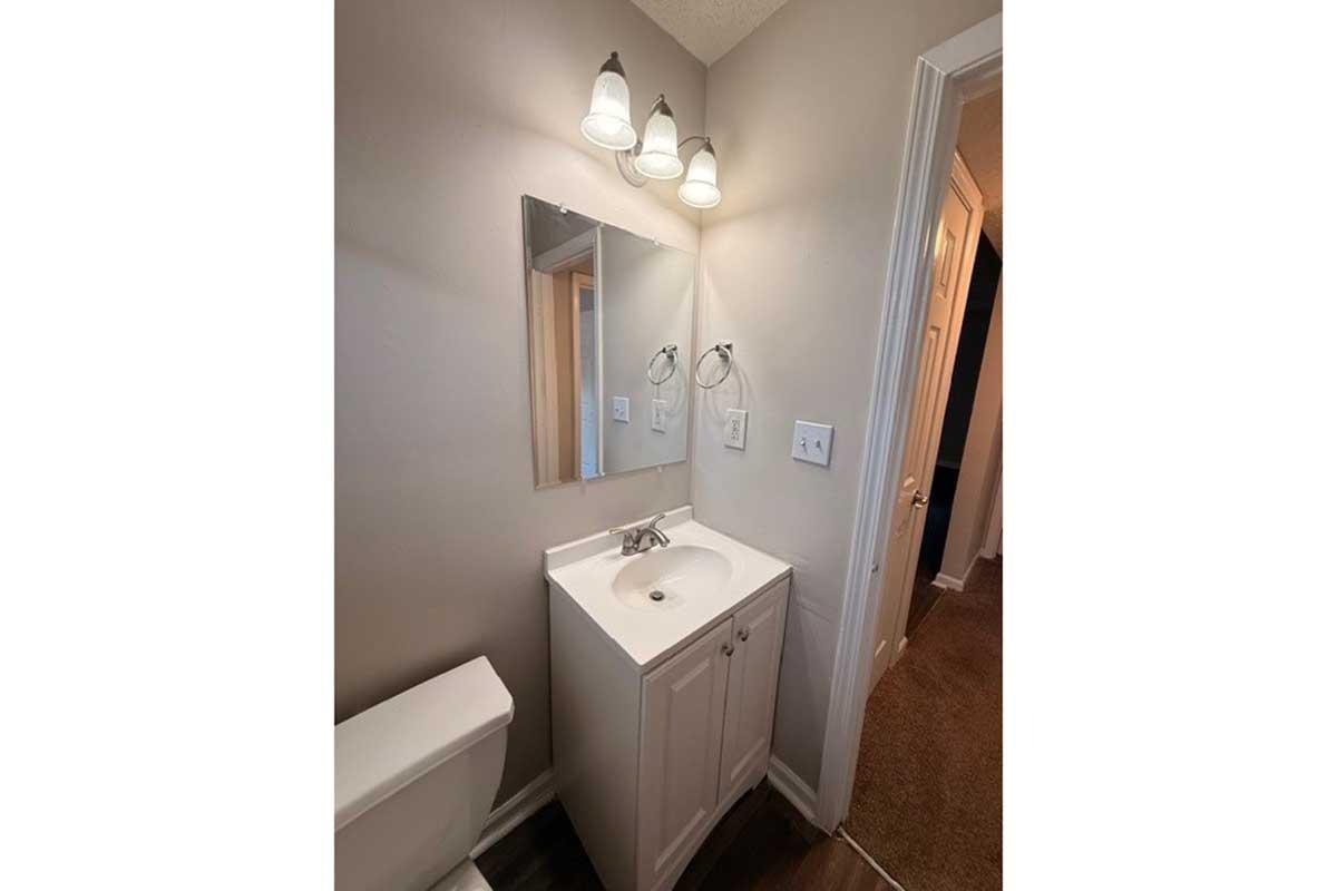 A small bathroom featuring a white vanity with a sink, a mirror above it, and two wall-mounted lights. A toilet is positioned next to the vanity. The walls are painted in a light color, and there is a doorway leading to another room. The floor has dark-colored flooring, contributing to a modern look.