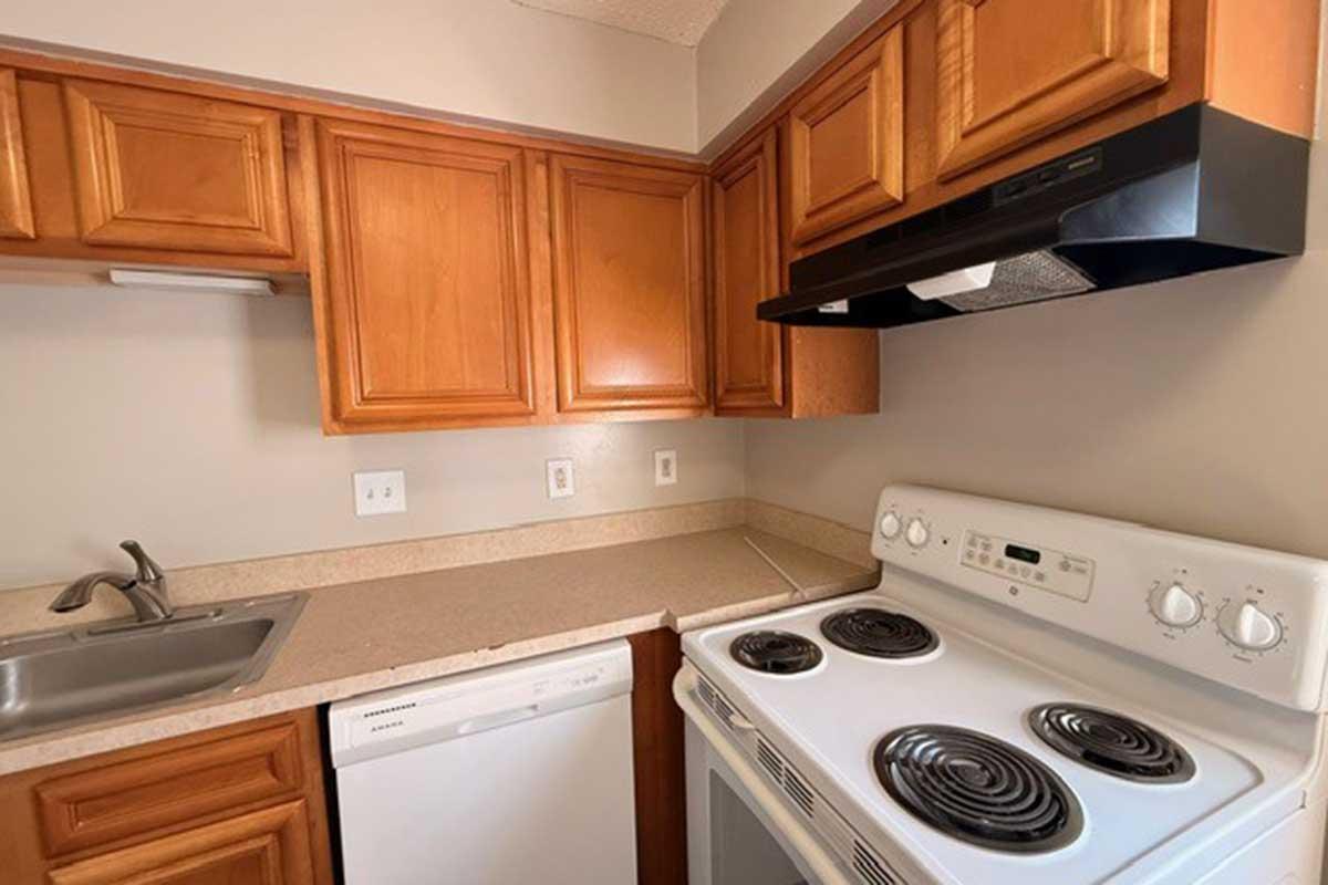 A modern kitchen featuring wooden cabinets, a stainless steel sink, a black range hood, a white stove with four burners, and a white dishwasher. The countertop is beige, and the walls are painted a light color, creating a clean and functional cooking space.