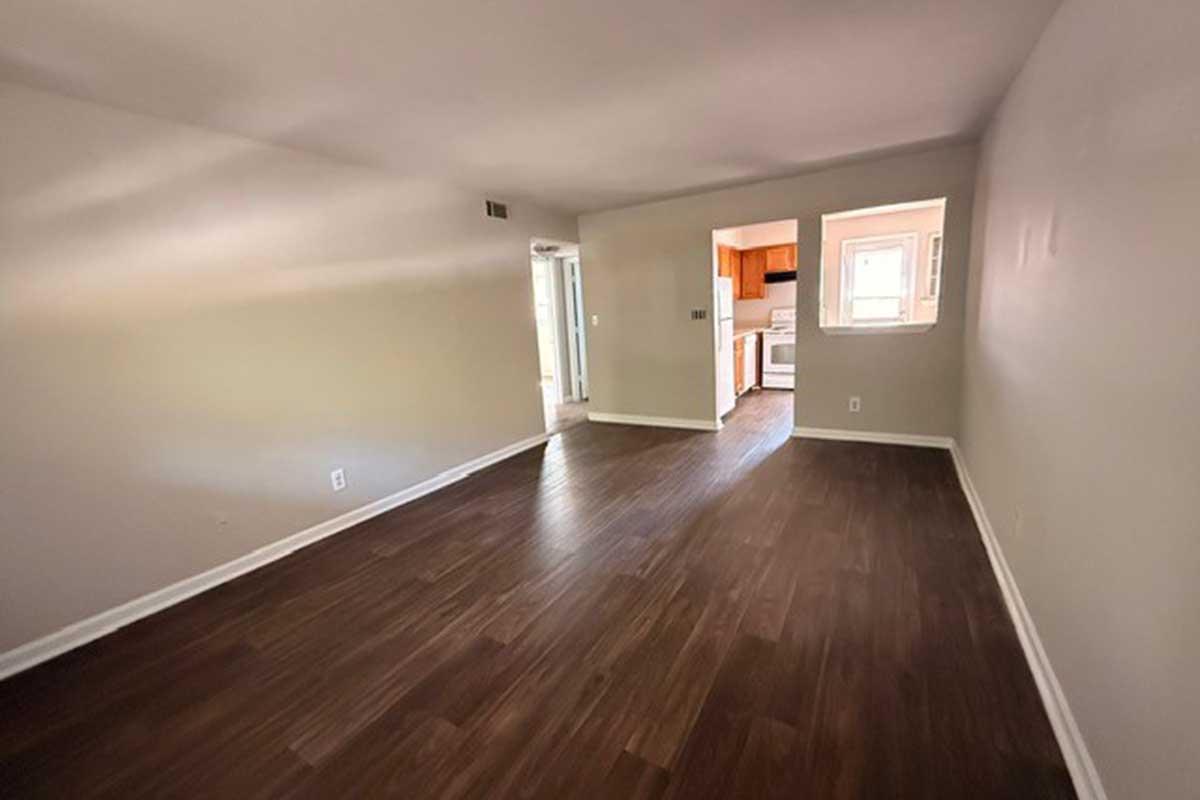 A spacious, empty room with light-colored walls and hardwood flooring. In the background, a doorway leads to a kitchen with wooden cabinetry. Sunlight illuminates the area, creating a bright, open atmosphere. The space is uncluttered, suitable for various furnishing options.