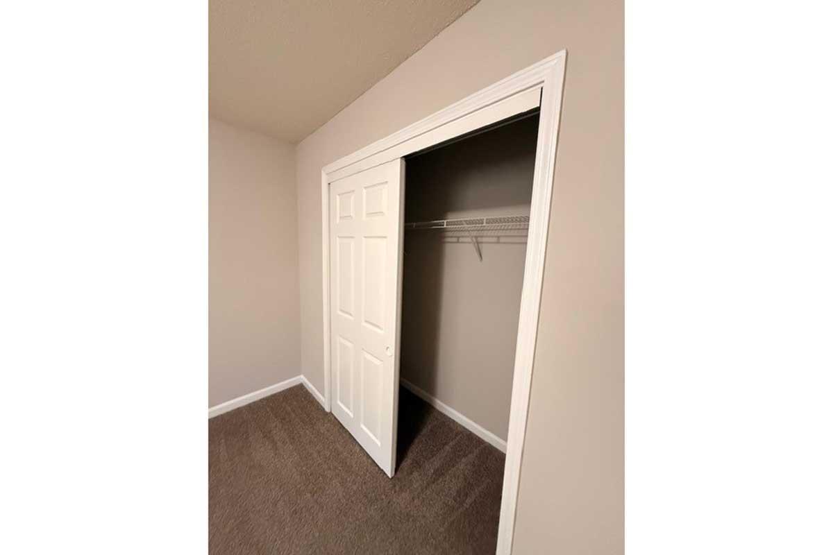 A view of a closet with a sliding white door partially open, revealing a simple interior with a metal hanging rod and beige walls. The floor is carpeted in soft brown, complementing the neutral color scheme of the room.