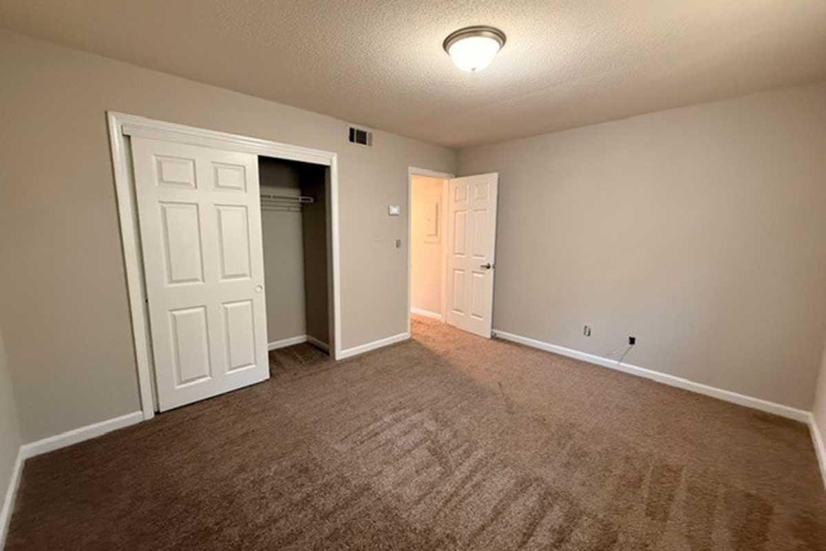 Empty room with beige walls and light brown carpet. There are two white doors, one leading to a closet and the other to an adjacent room. A ceiling light fixture is centrally located, and the space is well-lit, creating a clean and neutral atmosphere.