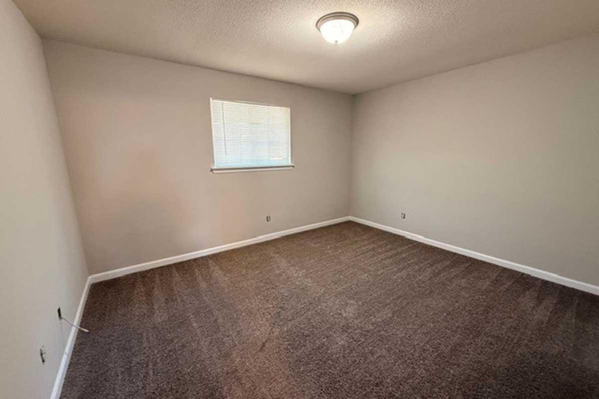 A clean, empty room with light gray walls and plush brown carpet. There is a window with white blinds allowing natural light in. The ceiling features a simple light fixture. The space appears well-maintained and is free from furniture or decorations.