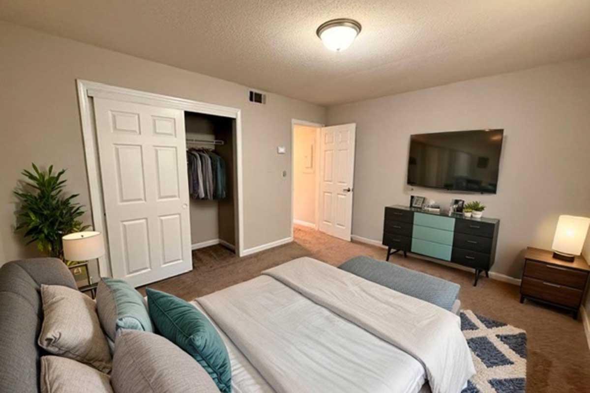 A cozy bedroom featuring a neatly made bed with decorative pillows, a stylish blue comforter, a television mounted on the wall, and a dresser. There is a closet with clothes visible, an armchair in the corner, and a bedside lamp. The walls are painted a soft beige, creating a warm atmosphere.