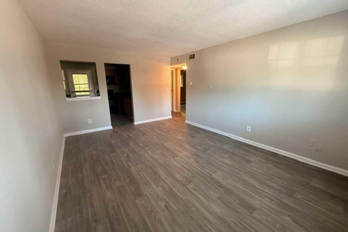 A spacious, empty room with light-colored walls and laminate flooring. There is a doorway leading to another room on the left and an open space that connects to a kitchen area in the background. The natural light enhances the room's brightness.