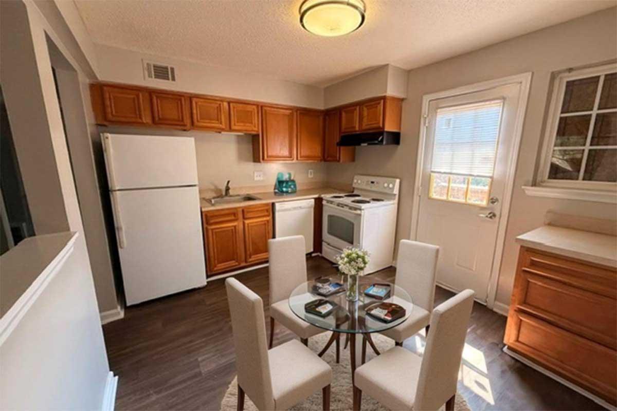 A modern kitchen with wooden cabinets and white appliances. There's a round glass dining table set with four light-colored chairs. A small vase with flowers is on the table, and sunlight is coming through a window. The kitchen features a fridge, stove, and sink against a neutral-colored wall.