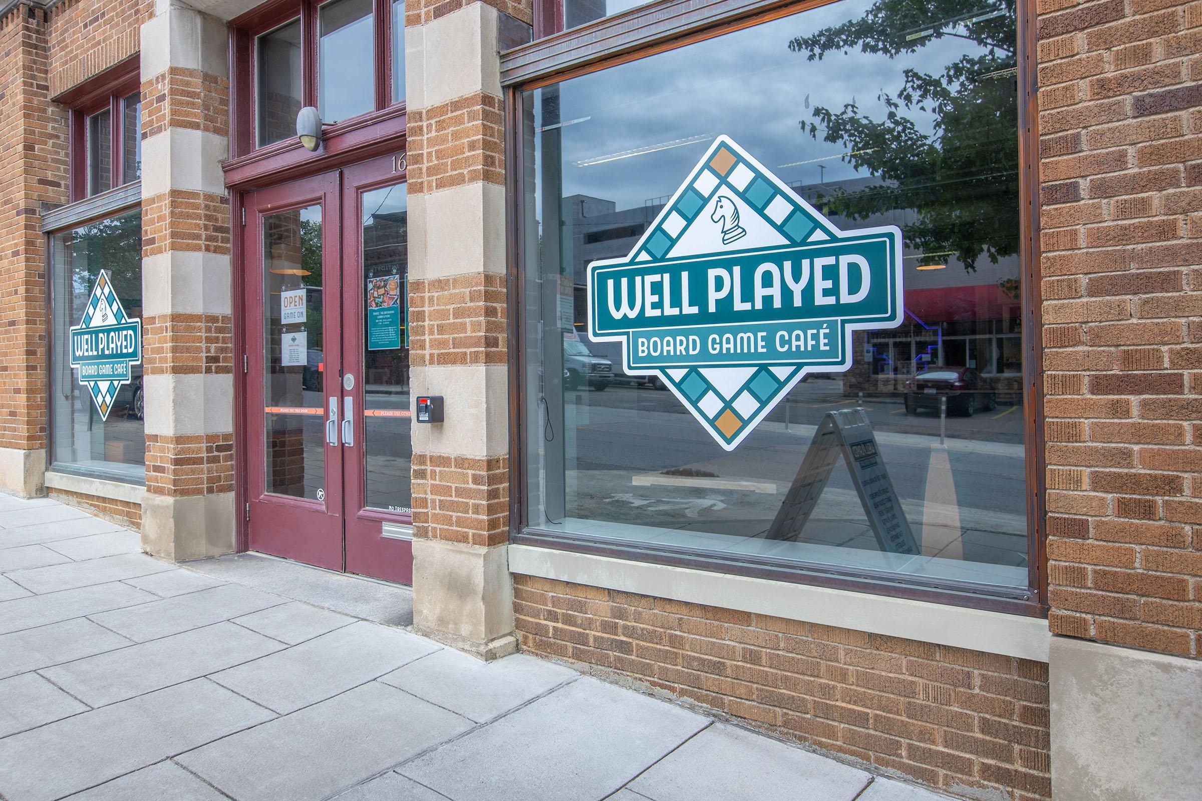 Exterior view of a board game café named "Well Played," featuring large glass windows with the café's logo displayed prominently. The building has a classic brick facade, and a sign with information about the café is placed in front. The area appears inviting for board game enthusiasts.