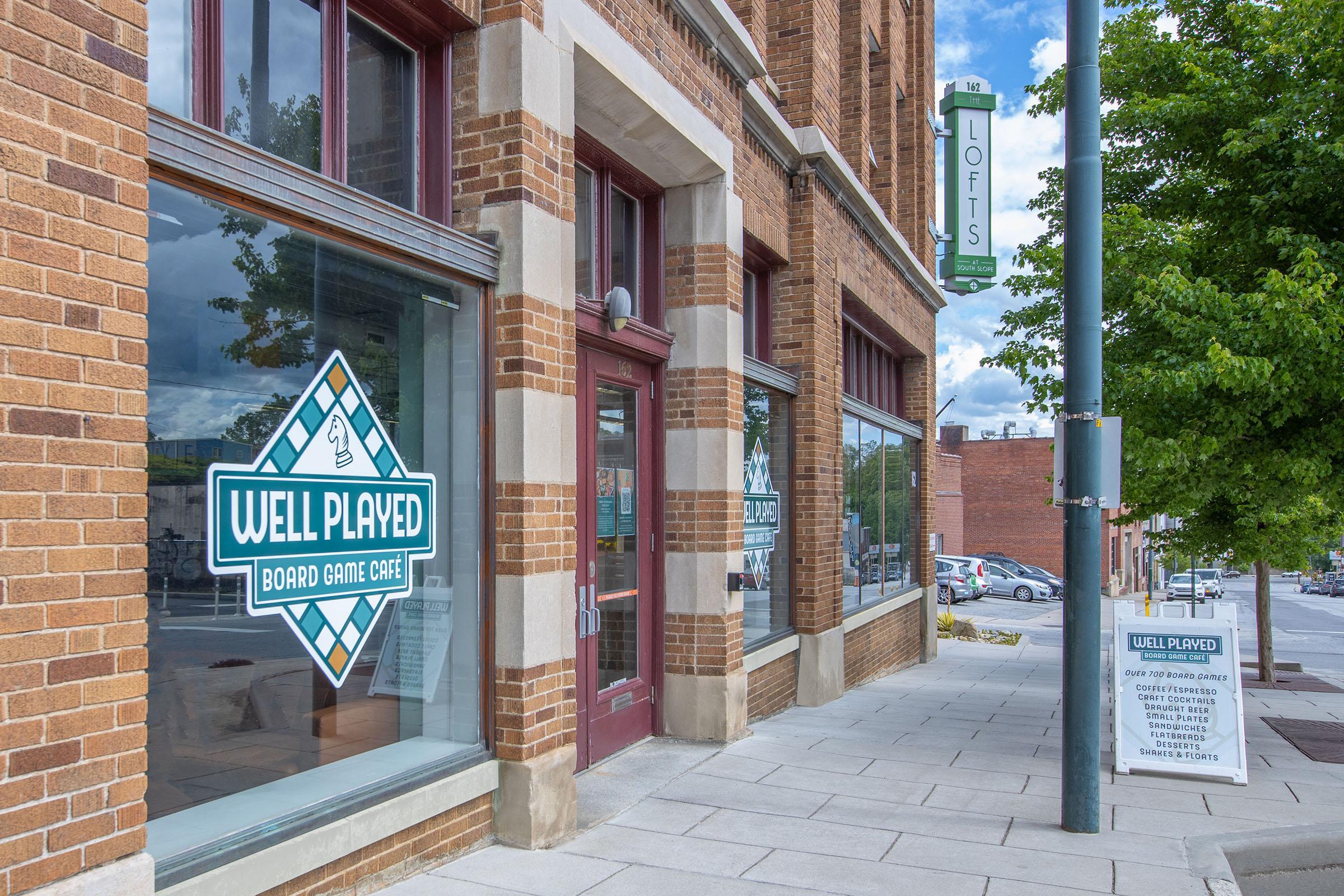 Exterior view of a board game cafe called "Well Played," featuring large windows with the cafe's logo. The building has a brick facade with decorative elements and is located in an urban area. Nearby signage indicates loft apartments, and the sidewalk is lined with parked cars and greenery.