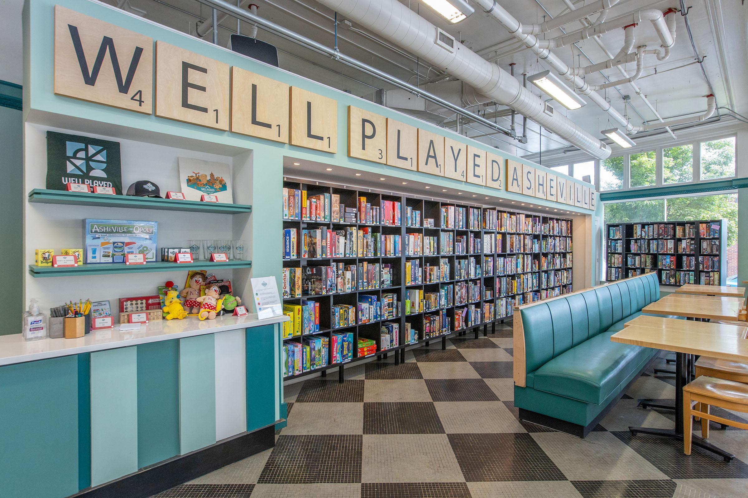 Interior of a board game café called "Well Played Asheville." Features a bright space with a large sign reading "WELL PLAYED ASHEVILLE," shelves filled with various board games, a seating area with teal booths, and a counter with colorful items, suggesting a welcoming environment for game enthusiasts.