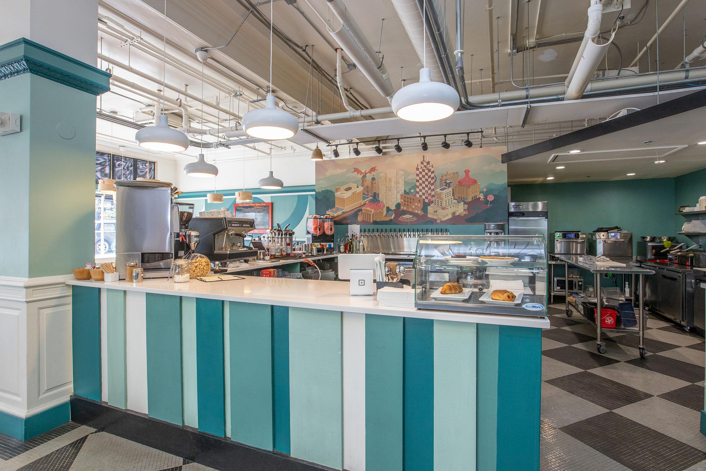 A modern café interior featuring a counter with pastries and drinks, flanked by shelves and kitchen equipment. The walls are adorned with a colorful mural depicting cityscapes. Bright lighting and a contemporary design create an inviting atmosphere. Black and white checkered flooring adds a retro touch.