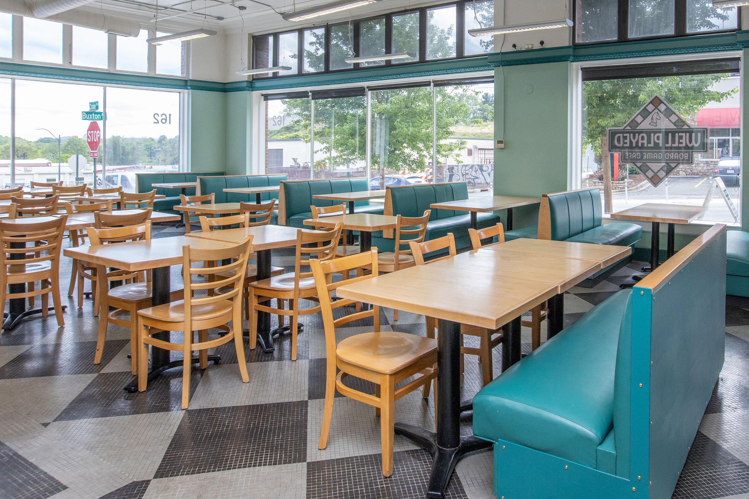 Interior of a casual dining restaurant featuring wooden tables and chairs, turquoise benches along the walls, large windows allowing natural light, and a checkered floor. The space is clean and inviting, with a view of the outside.
