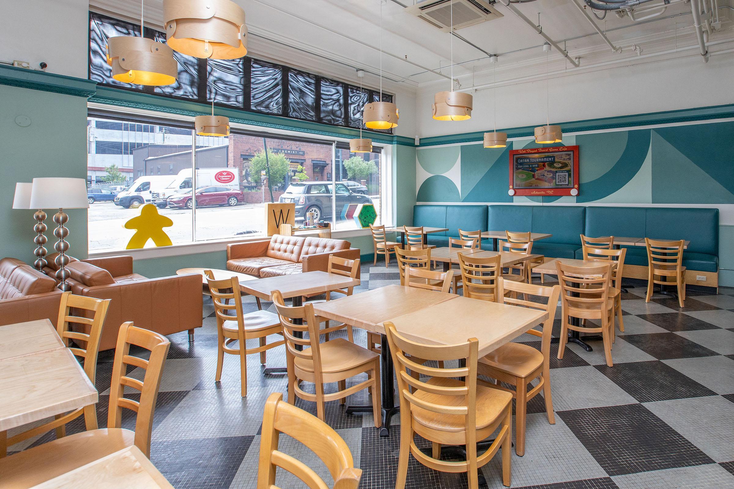 Interior of a modern café featuring wooden tables and chairs, a brown sofa, and decorative wall art with geometric patterns. Large windows provide natural light, and there are several pendant lights hanging from the ceiling. A playful yellow figure is visible in the window.