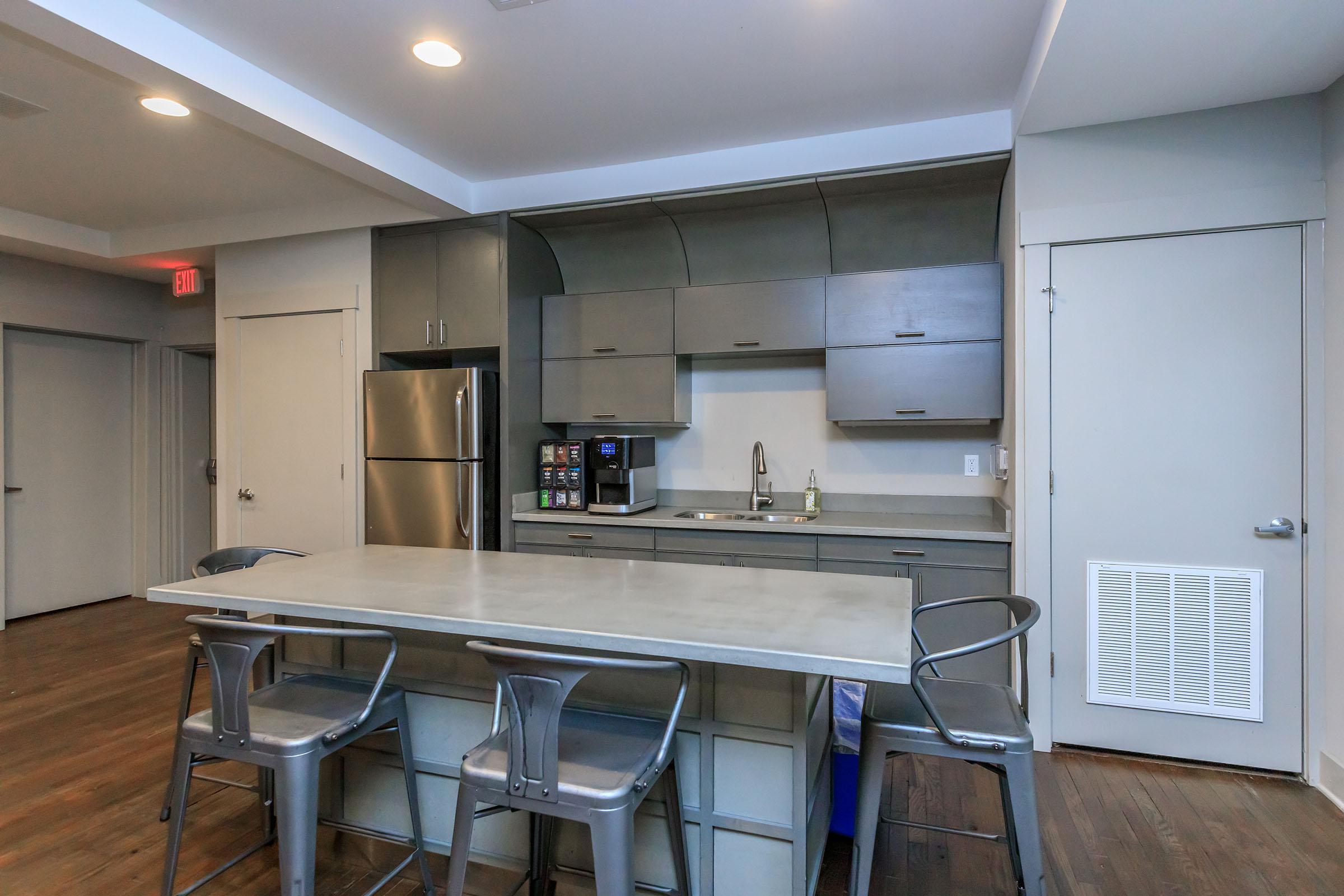 Modern kitchen featuring a gray cabinetry setup with a stainless steel refrigerator, a sink, and a coffee maker. A large concrete island with metal stools provides seating. The space is well-lit with recessed lighting and has a clean, minimalist design. Hardwood floors add warmth to the contemporary aesthetic.