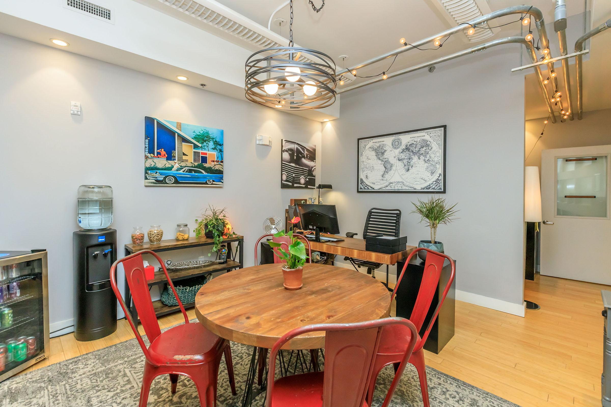 A stylish office space with a round wooden table surrounded by red chairs. The walls feature framed artwork, including a vintage car painting and a world map. There's a water cooler and a small table with jars, while plants add a touch of greenery. Modern lighting fixtures enhance the inviting atmosphere.