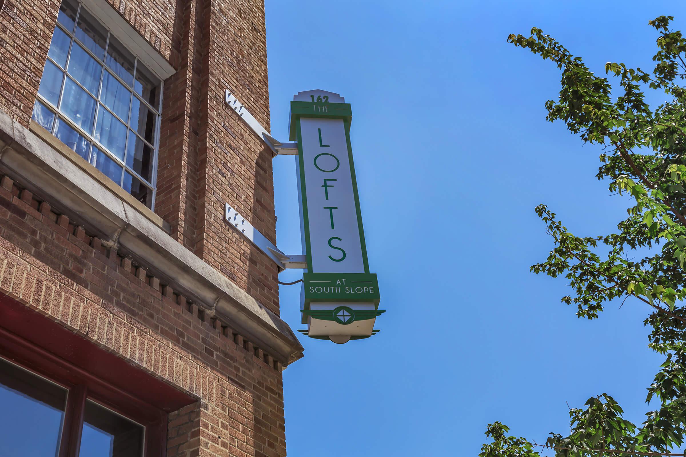 Sign for "Lofts at South Slope" mounted on a brick building, featuring a green and white design with the word "LOFTS" prominently displayed. Clear blue sky in the background, with some greenery visible from a nearby tree.