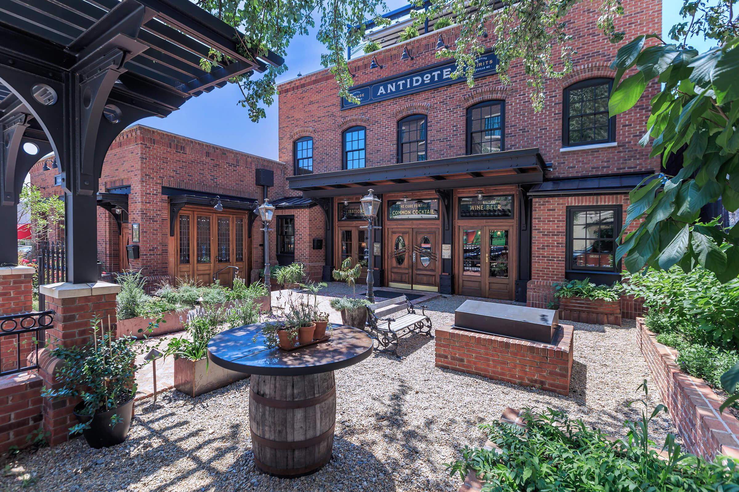 A charming outdoor patio area featuring a wooden table with potted plants, surrounded by lush greenery and gravel pathways. The backdrop includes a historic brick building with wooden doors and the sign "ANTIDOTE." Bright blue sky enhances the inviting atmosphere of the space.
