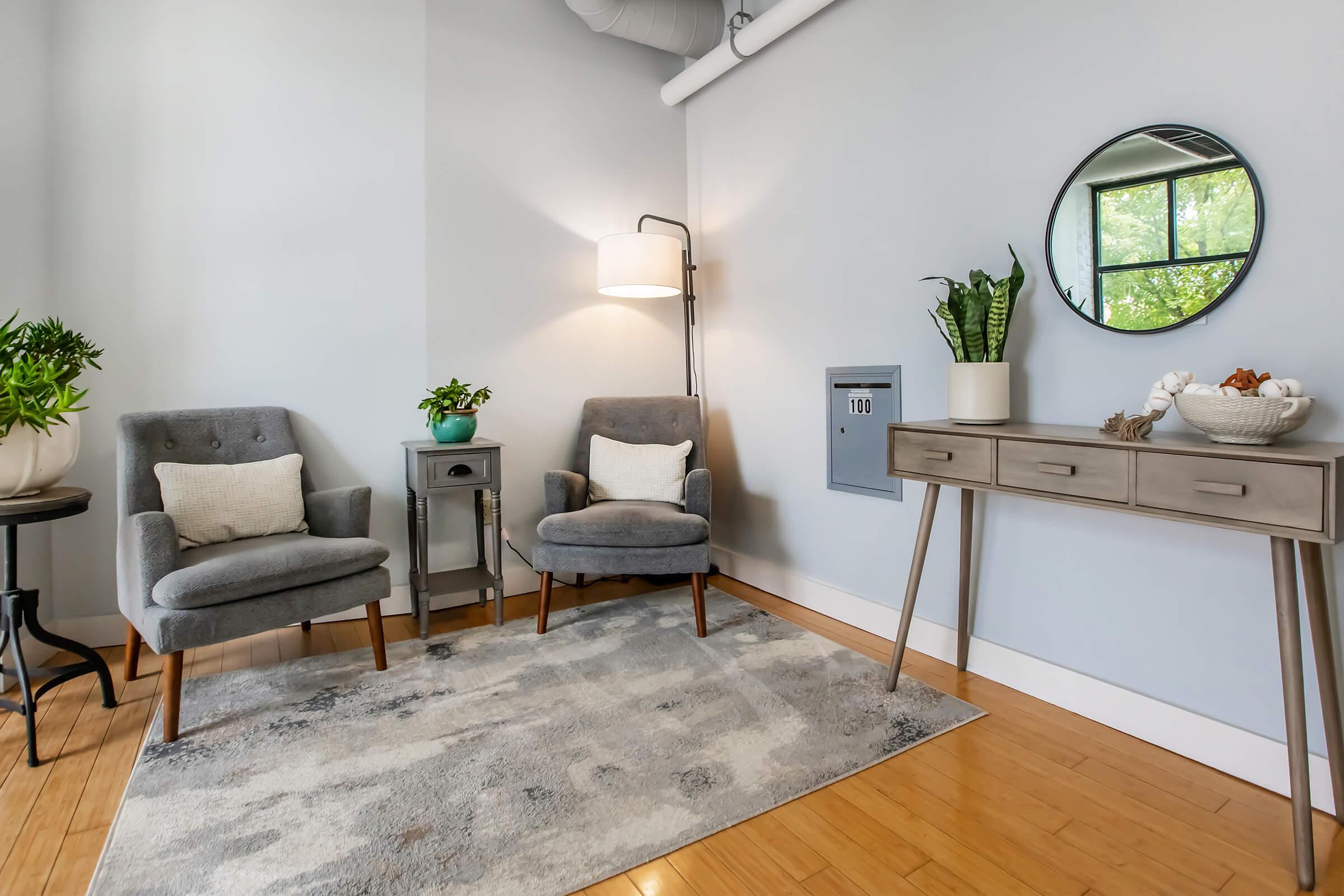 A cozy living space featuring two gray upholstered chairs, a round mirror on the wall, a small side table with a plant, and a stylish console table adorned with decorative items and a potted plant. A floor lamp provides warm light, and a soft area rug adds comfort. Natural light enters through the window.