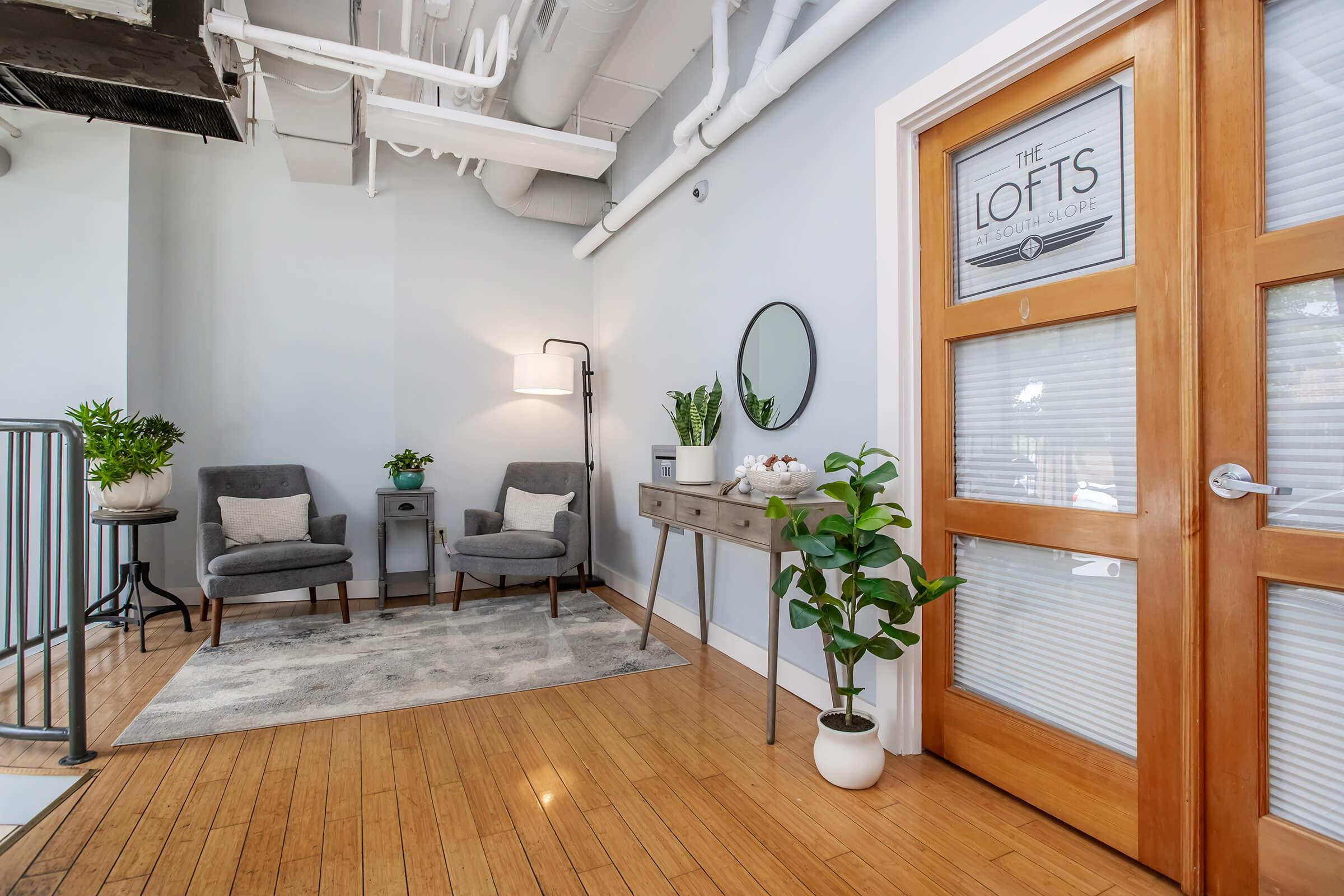 A cozy reception area featuring two gray armchairs, a small side table with a lamp, and a mirror. A decorative plant sits near the door, which has a sign reading "The Lofts." The space has wooden flooring and light blue walls, creating a welcoming environment. Large windows provide natural light.
