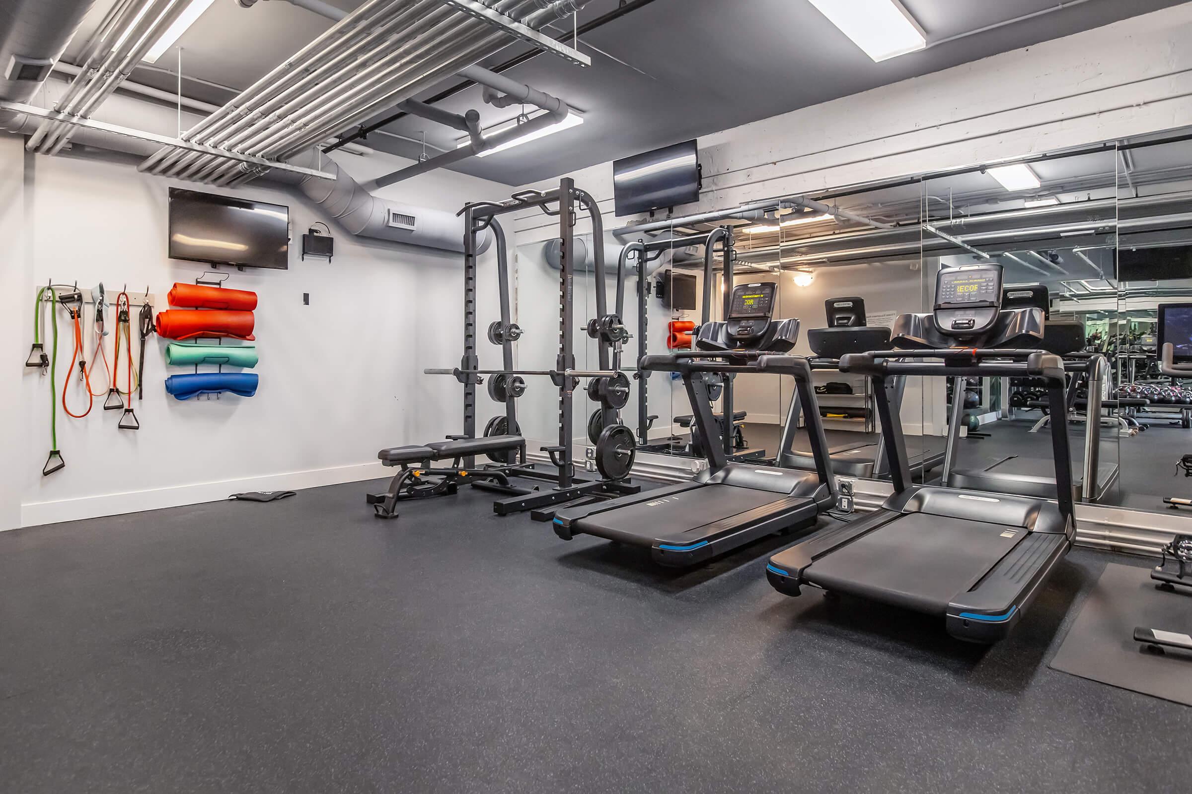 A modern gym interior featuring two treadmills, a weightlifting bench, and a multi-functional weight training machine. Colorful exercise mats are neatly stacked on the wall, and two flat-screen TVs are mounted on the ceiling. The flooring is dark rubber, and large mirrors create a spacious feel.