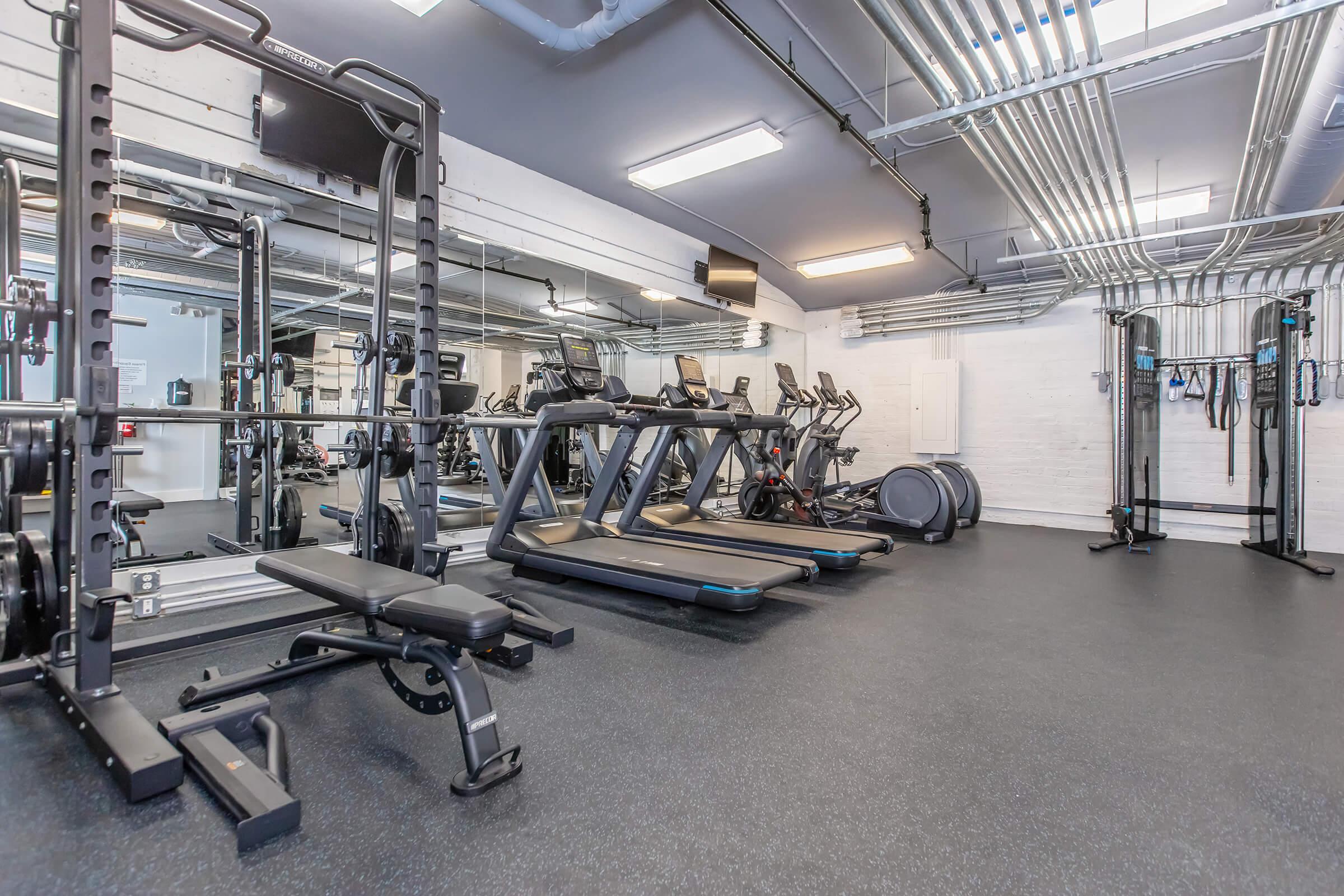 A modern gym interior featuring exercise equipment, including treadmills, weight benches, and resistance training machines. The space has large mirrors, industrial-style ceilings with visible pipes, and a clean, organized layout, creating an inviting atmosphere for workouts.