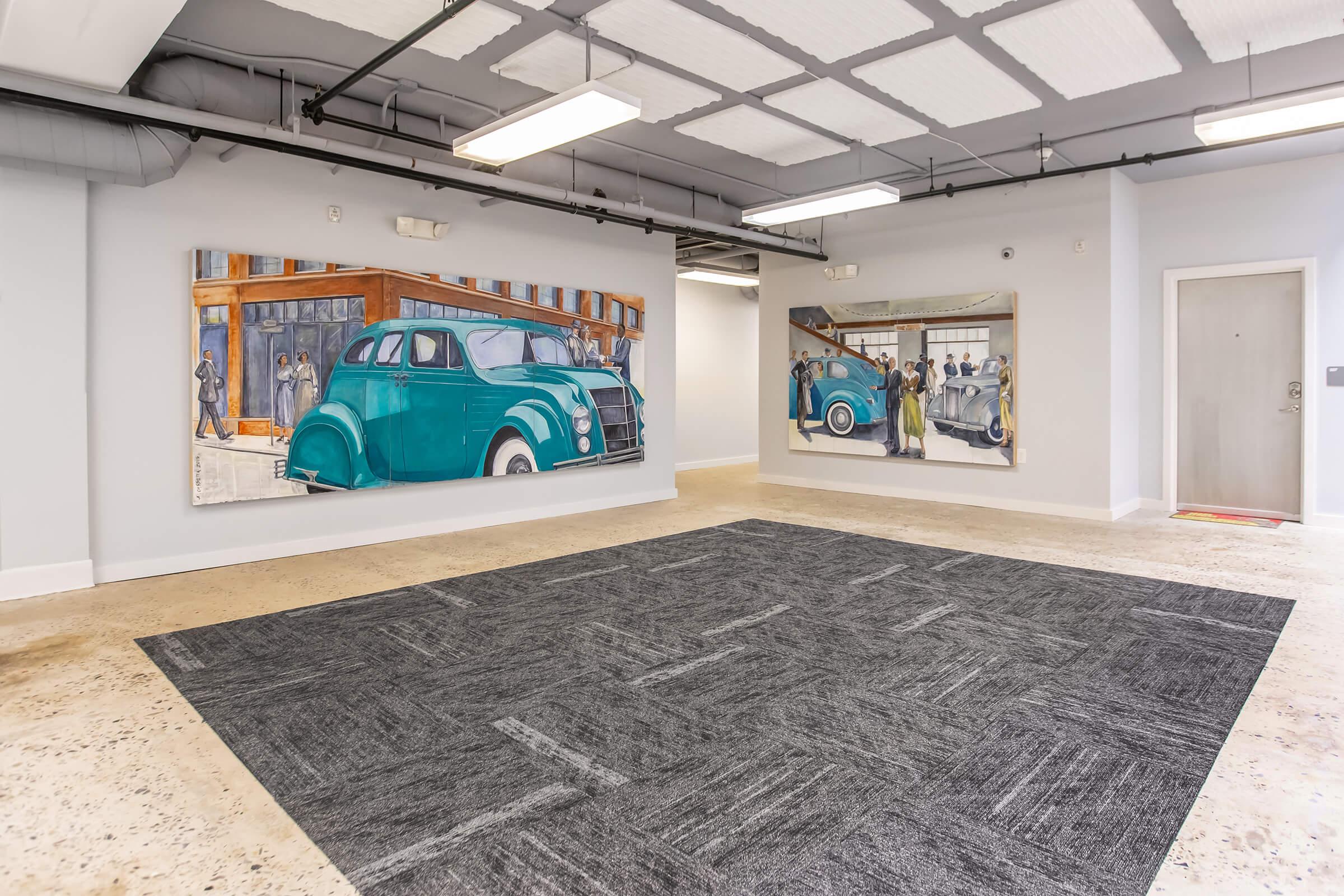 A spacious room with a textured carpet and modern industrial design. Two large murals on the walls depict vintage cars and scenes featuring people, adding artistic flair to the space. The room is well-lit with overhead lights and features a neutral color palette. A doorway leads to another area.