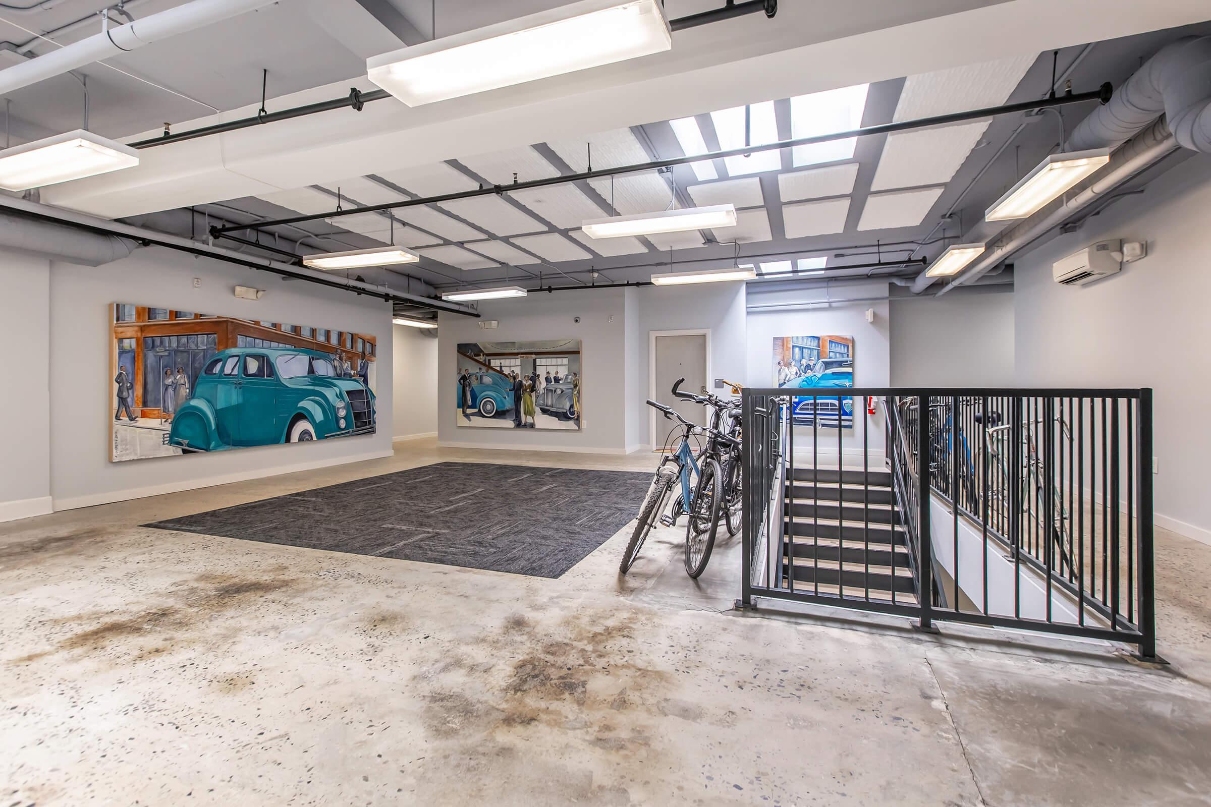 A spacious indoor area with polished concrete flooring, featuring two large wall murals of vintage cars. Bicycle racks are visible, and a staircase leads to another level. Bright overhead lighting illuminates the space, which has an open and modern design.