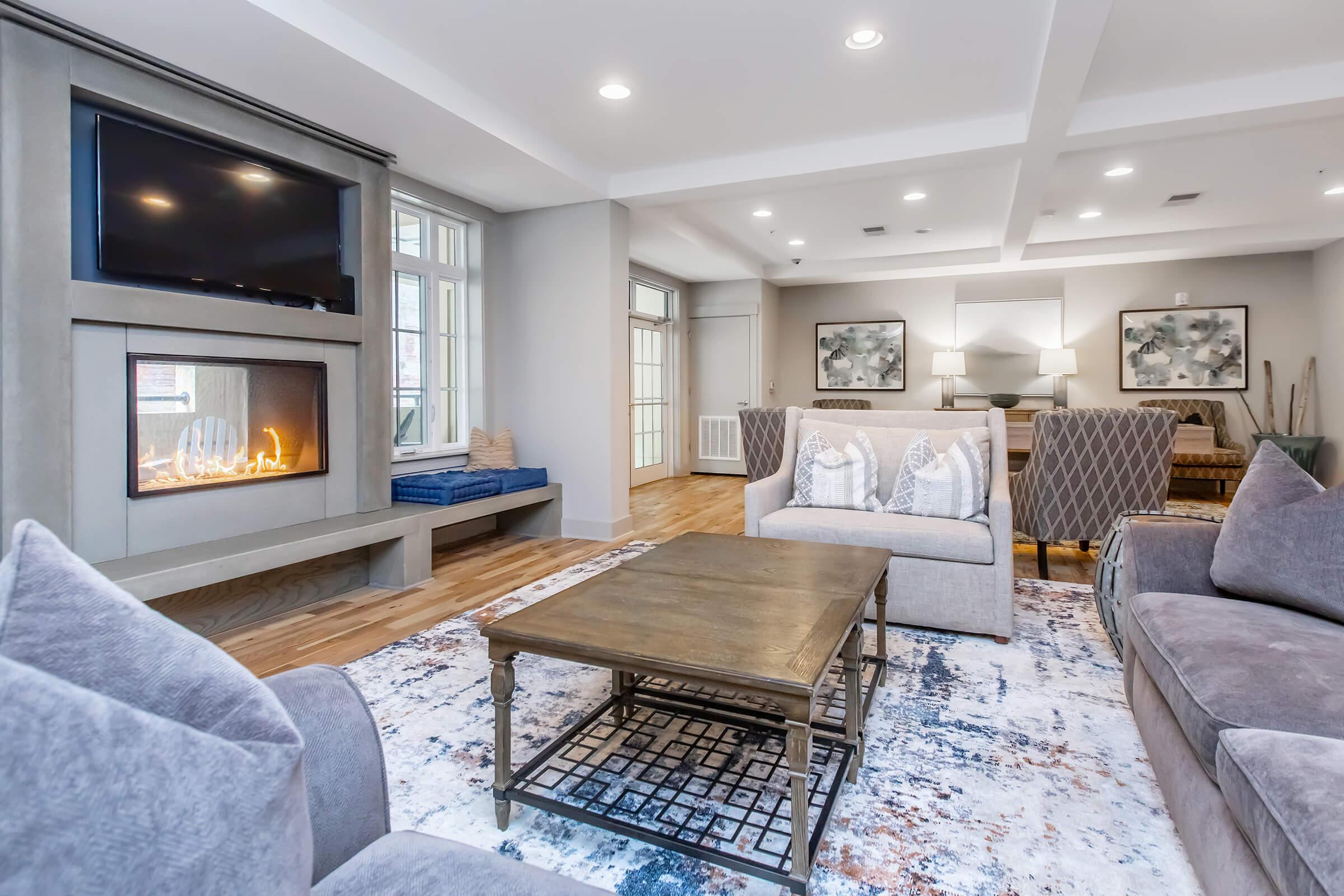A cozy living room featuring a modern design with a fireplace integrated into a wall. A plush sofa is complemented by decorative cushions, and a wooden coffee table sits on a patterned area rug. Large windows allow natural light, and a pair of stylish armchairs add elegance to the space.