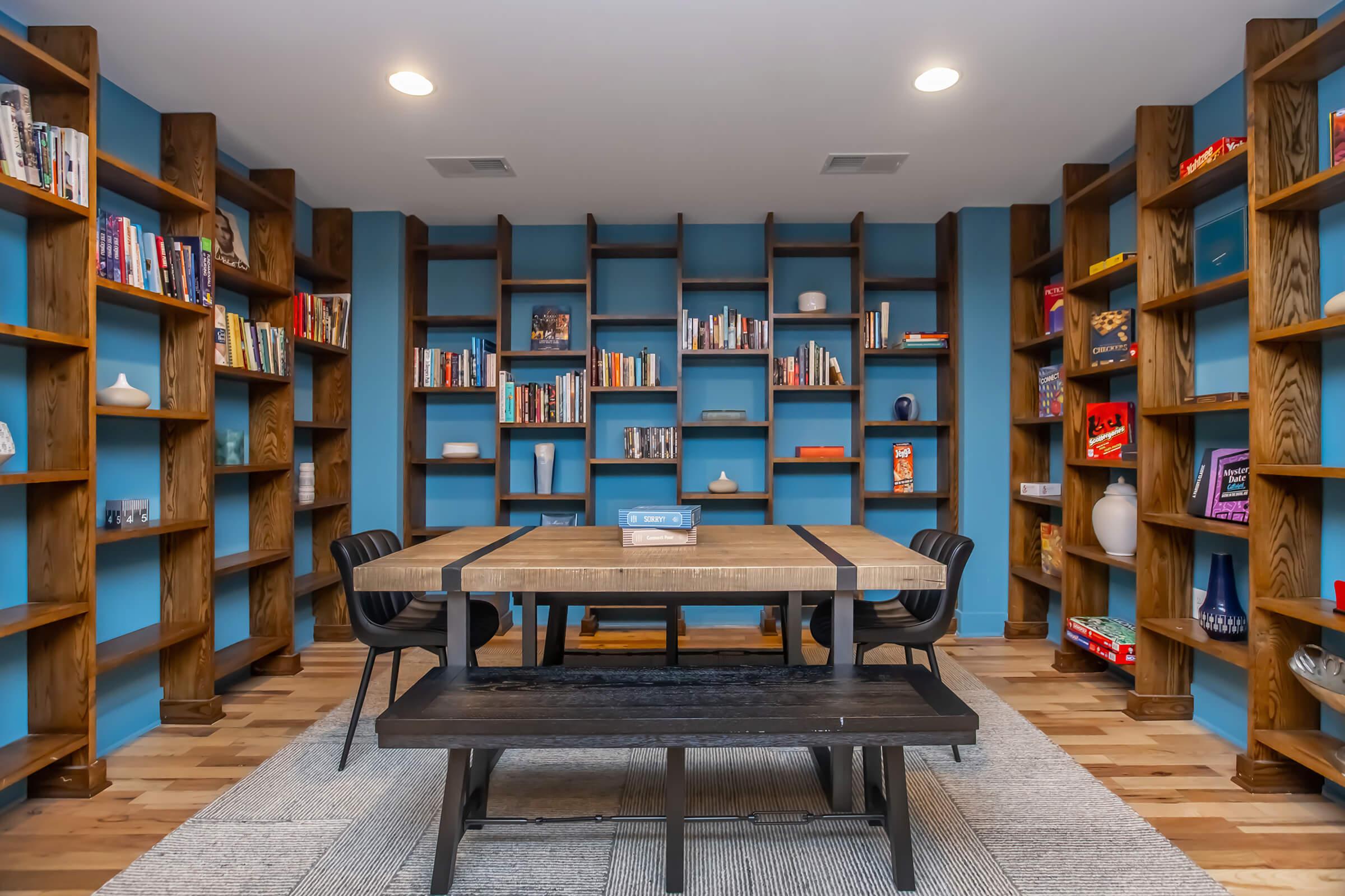 A cozy room featuring a wooden table surrounded by black chairs, with a textured rug beneath. The walls are painted a vibrant blue, and bookshelves filled with various books and decorative items line the walls. Soft lighting illuminates the space, creating an inviting atmosphere.